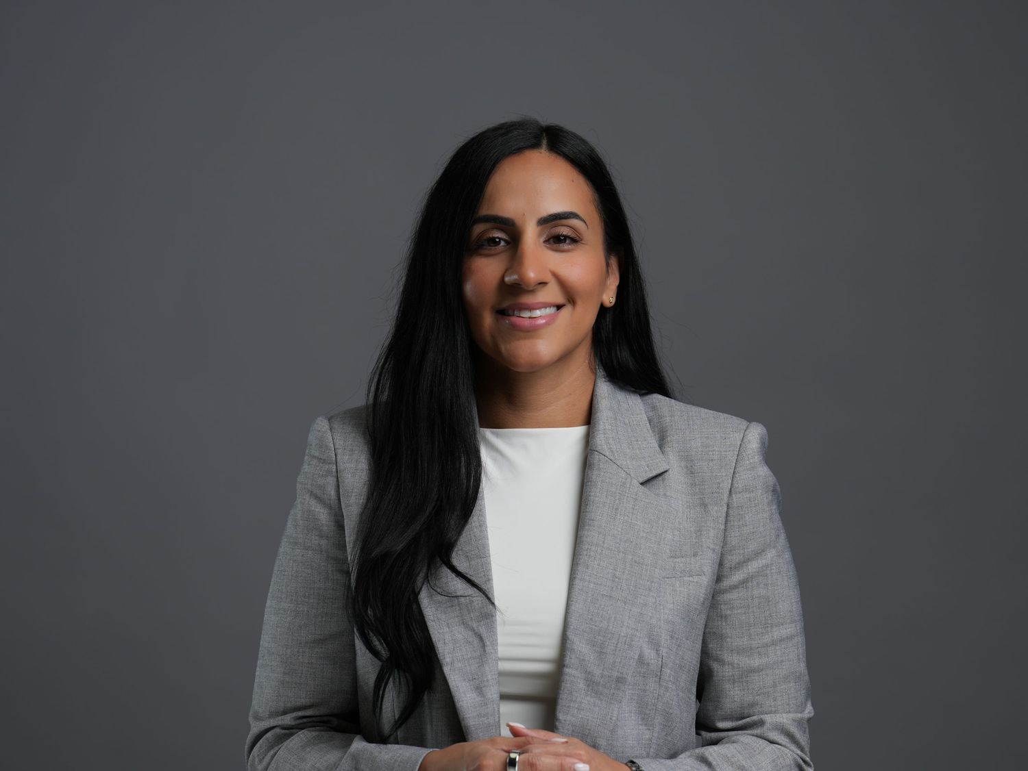 Professional woman in gray blazer smiling confidently with long dark hair against a gray background.
