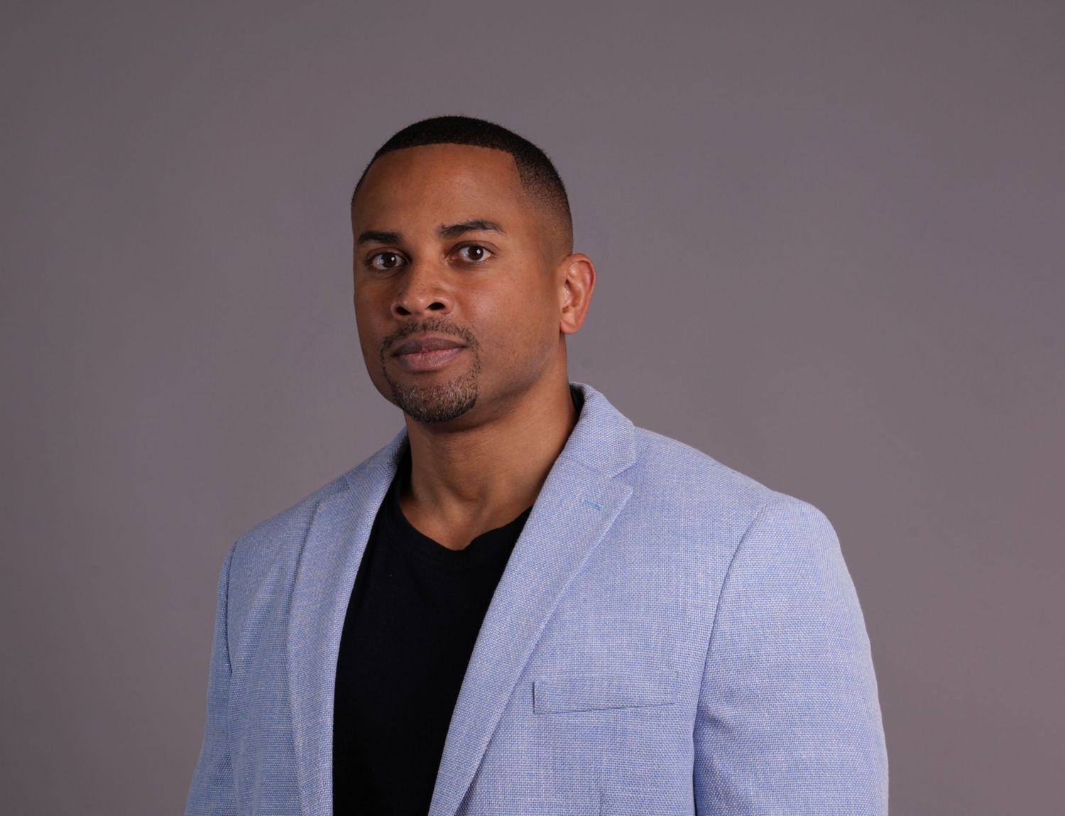 Professional man in a light blue blazer posing against a gray studio background with a confident expression.