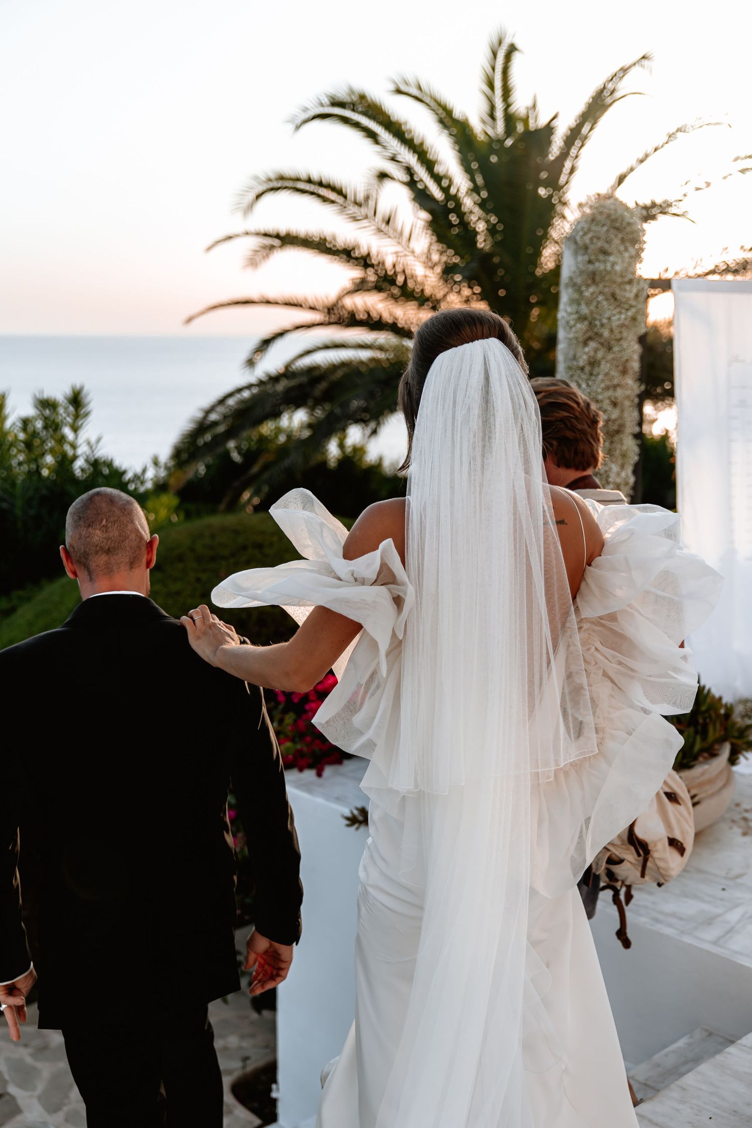A bride in a flowing white gown and veil walks up outdoor steps near a palm tree at sunset.