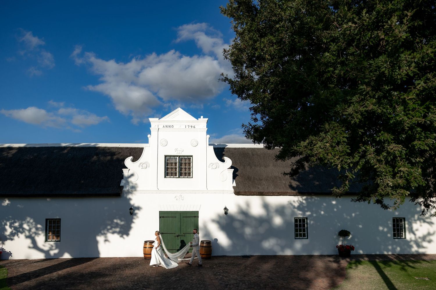 Historic Cape Dutch colonial architecture with white-washed walls and thatched roofs set against lush green trees and blue sky.