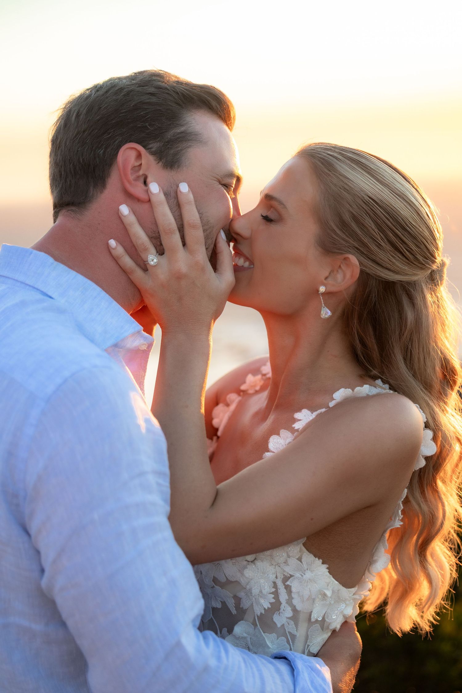 Bride and groom share a romantic sunset kiss, bride wearing white floral dress and diamond ring.