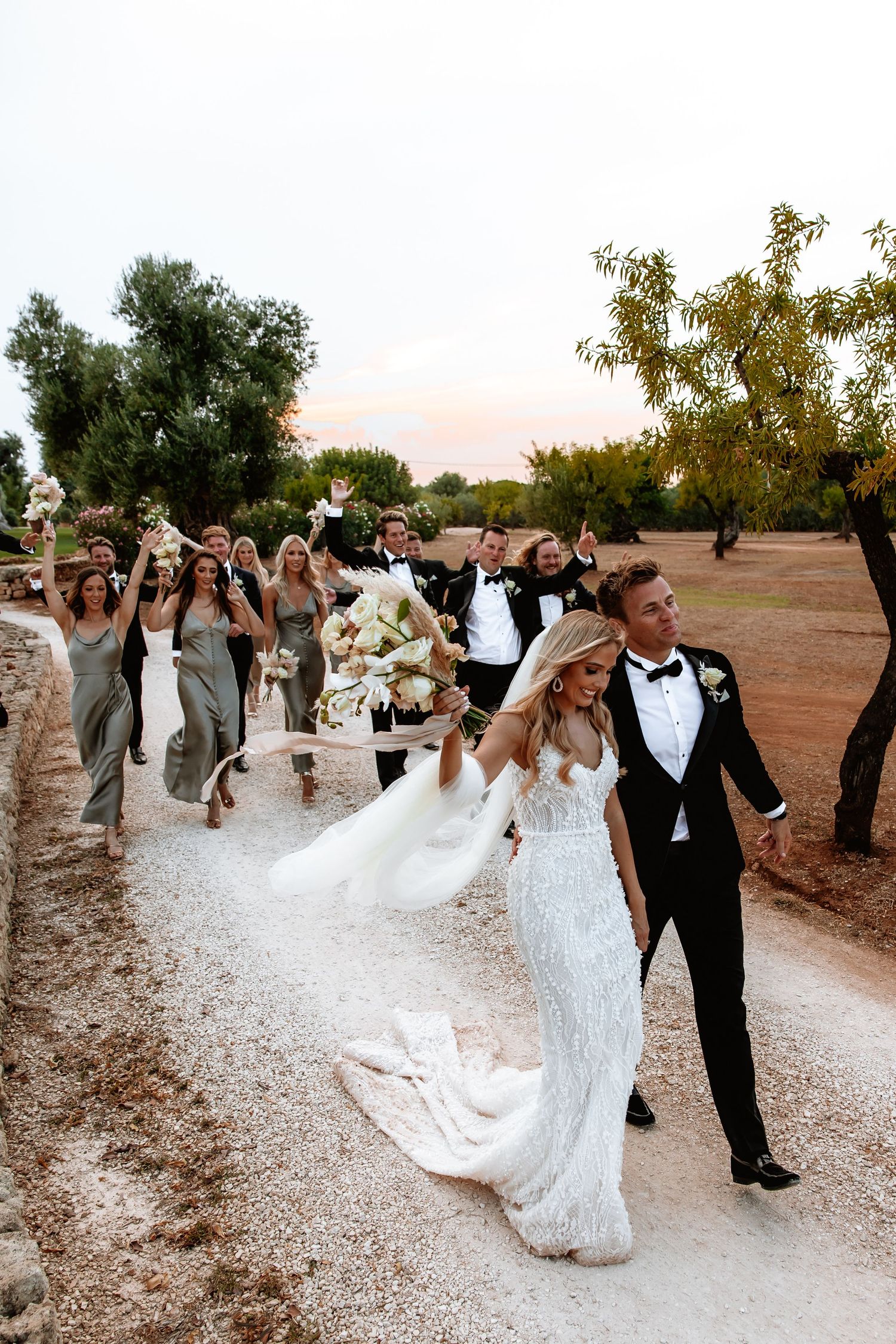 Wedding party walking and celebrating on a white gravel path through an olive grove.