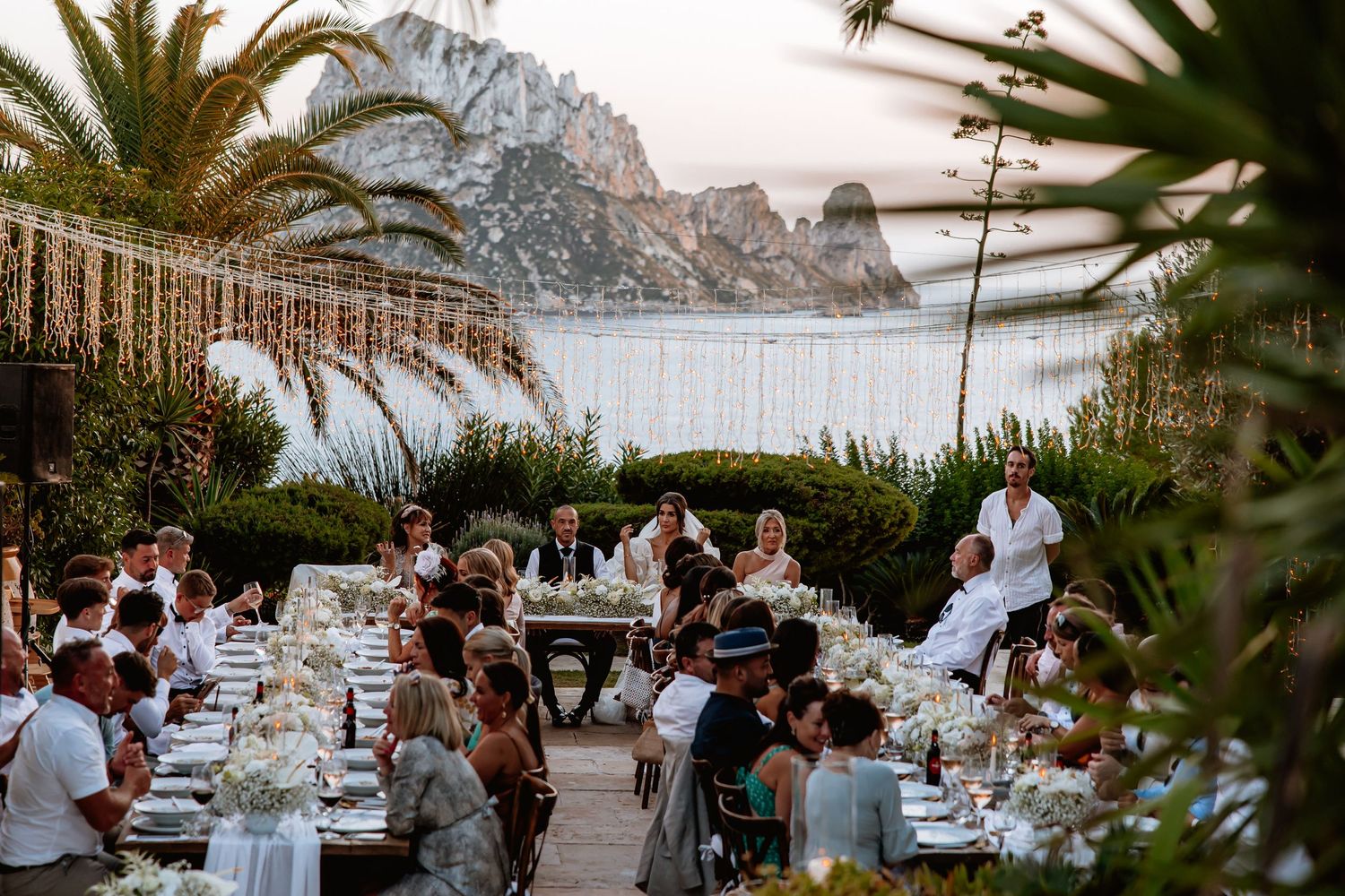 Wedding guests enjoy an elegant outdoor reception dinner at sunset with stunning coastal views and palm trees.