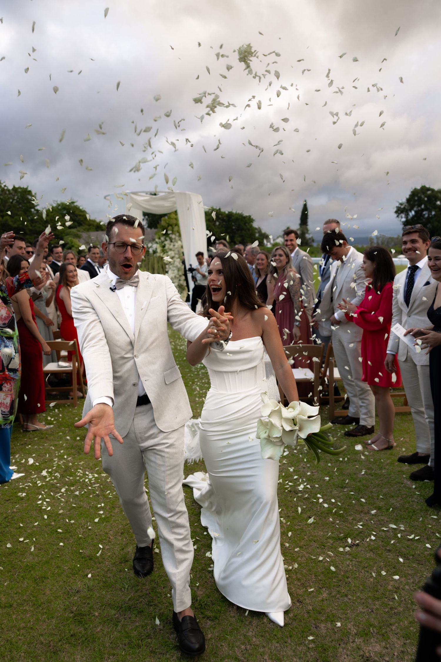 Wedding guests shower a couple with white flower petals during an outdoor celebration as they walk down the aisle.