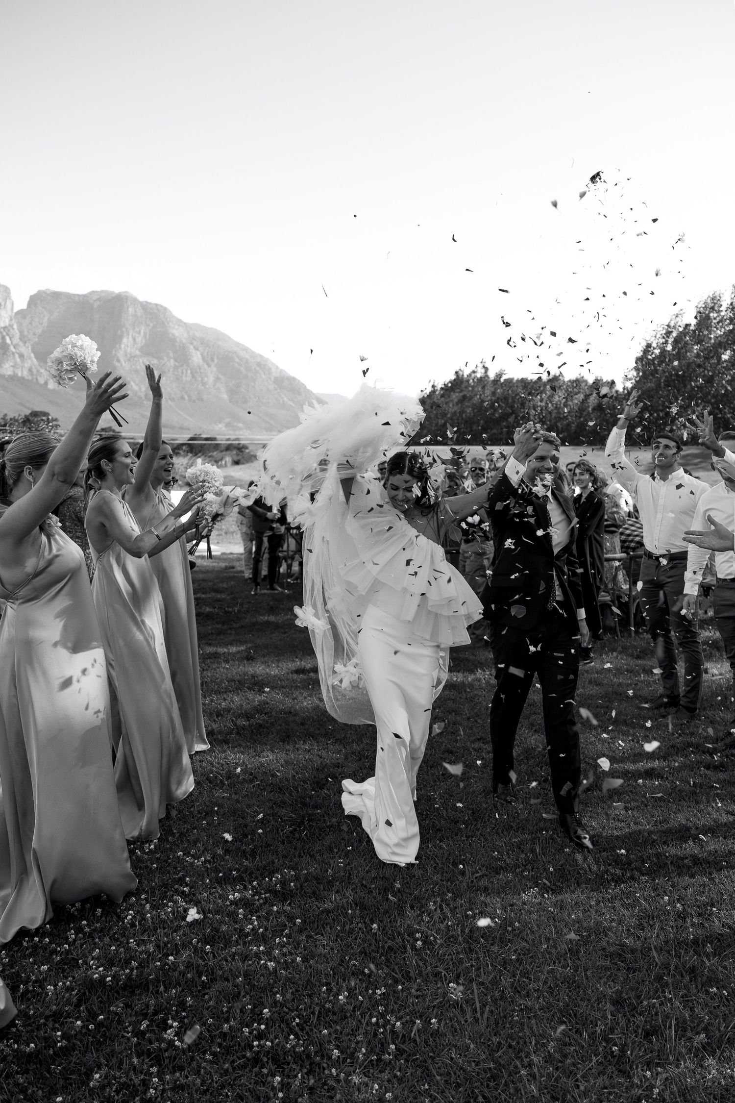 Wedding guests in white attire throw confetti and celebrate during an outdoor ceremony.