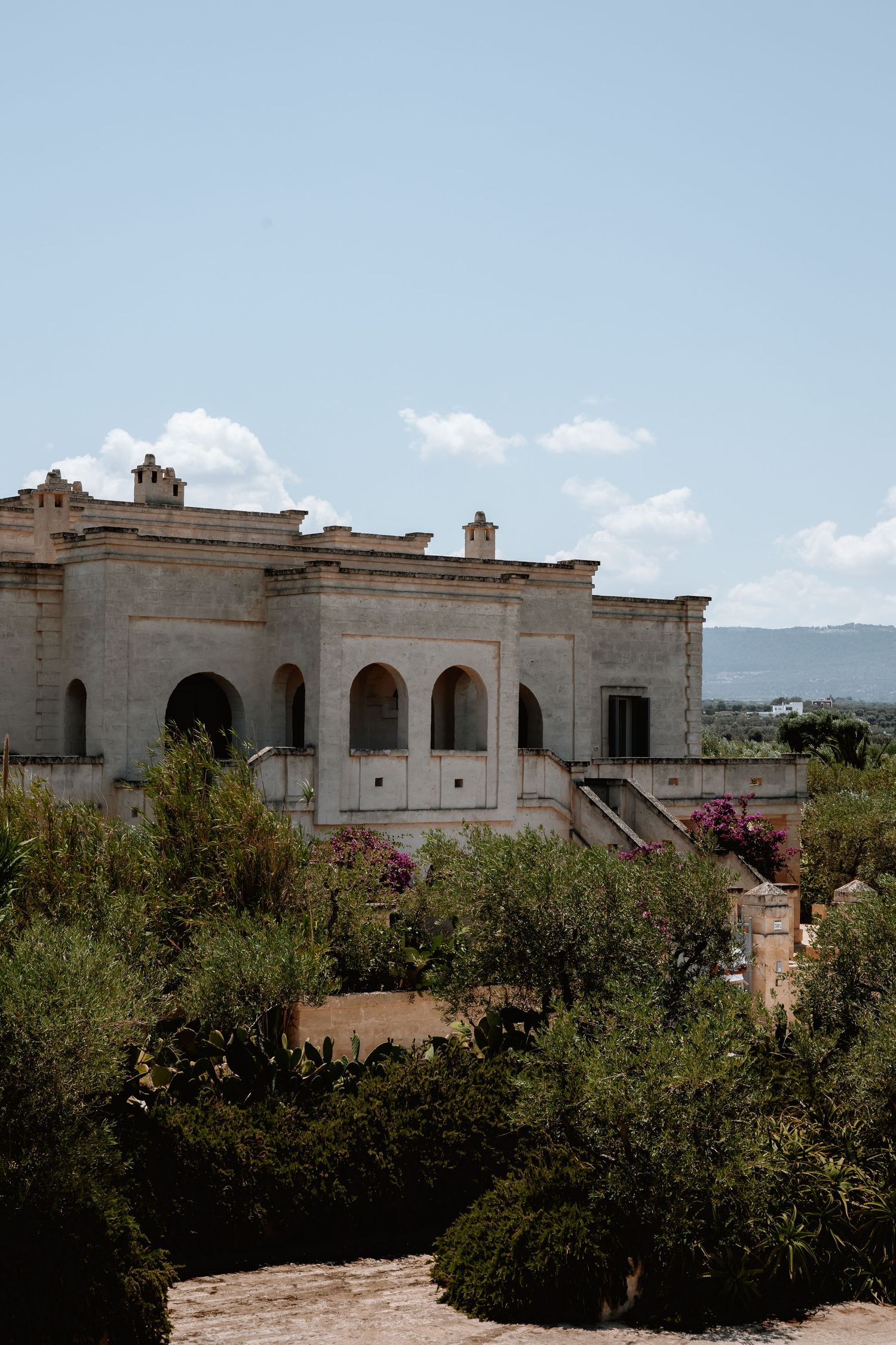 Elegant Mediterranean villa with white stone facade and arched windows surrounded by lush gardens and blue sky.
