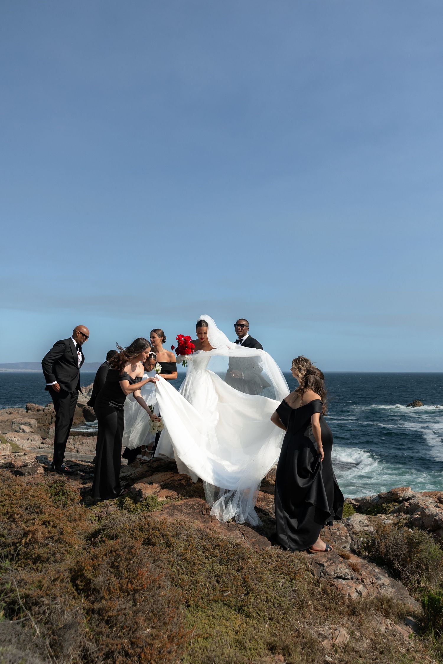 Wedding party on rocky oceanside cliff, bride in white gown with flowing veil, attendants in black assisting.