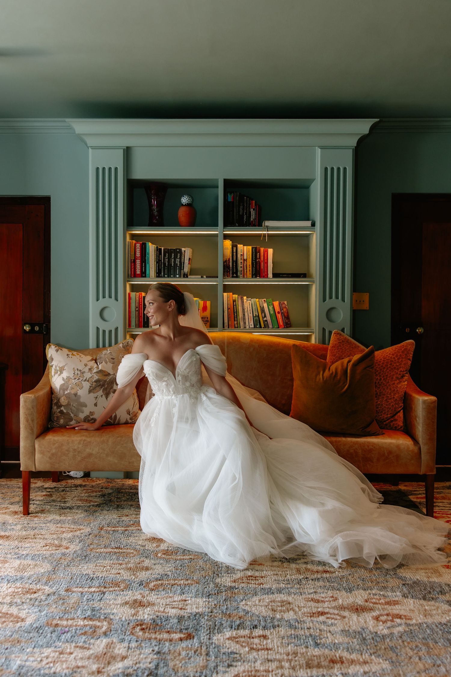 A person in a flowing white wedding gown reclines gracefully on an ornate rug in front of built-in bookshelves.