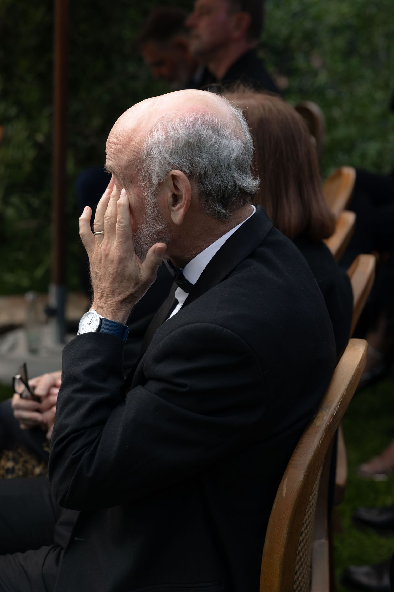 Sequence of images showing an elderly person in formal attire covering their face while seated in a wooden chair outdoors.