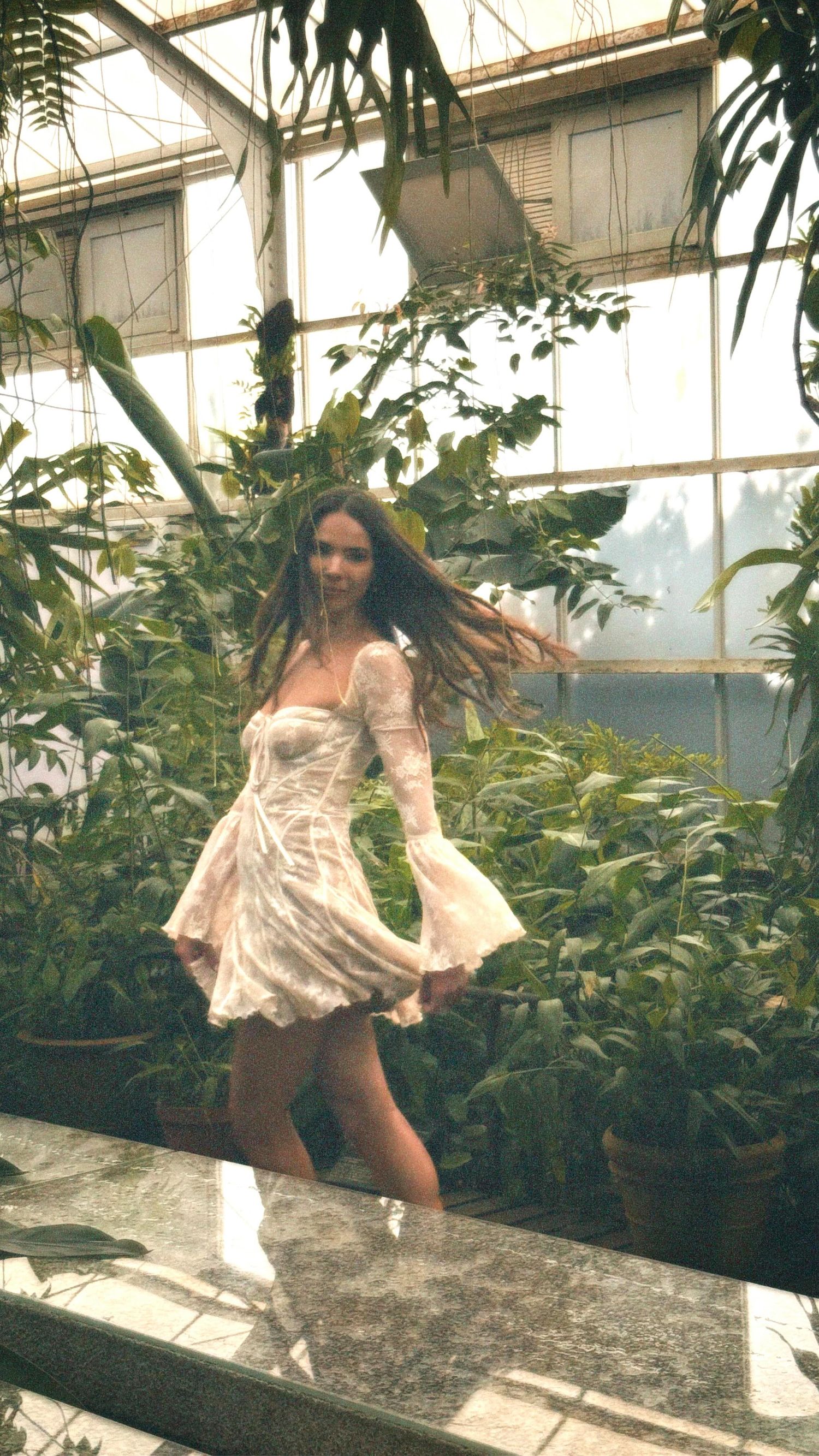 A person in a white dress poses playfully in a greenhouse surrounded by lush green plants and foliage.