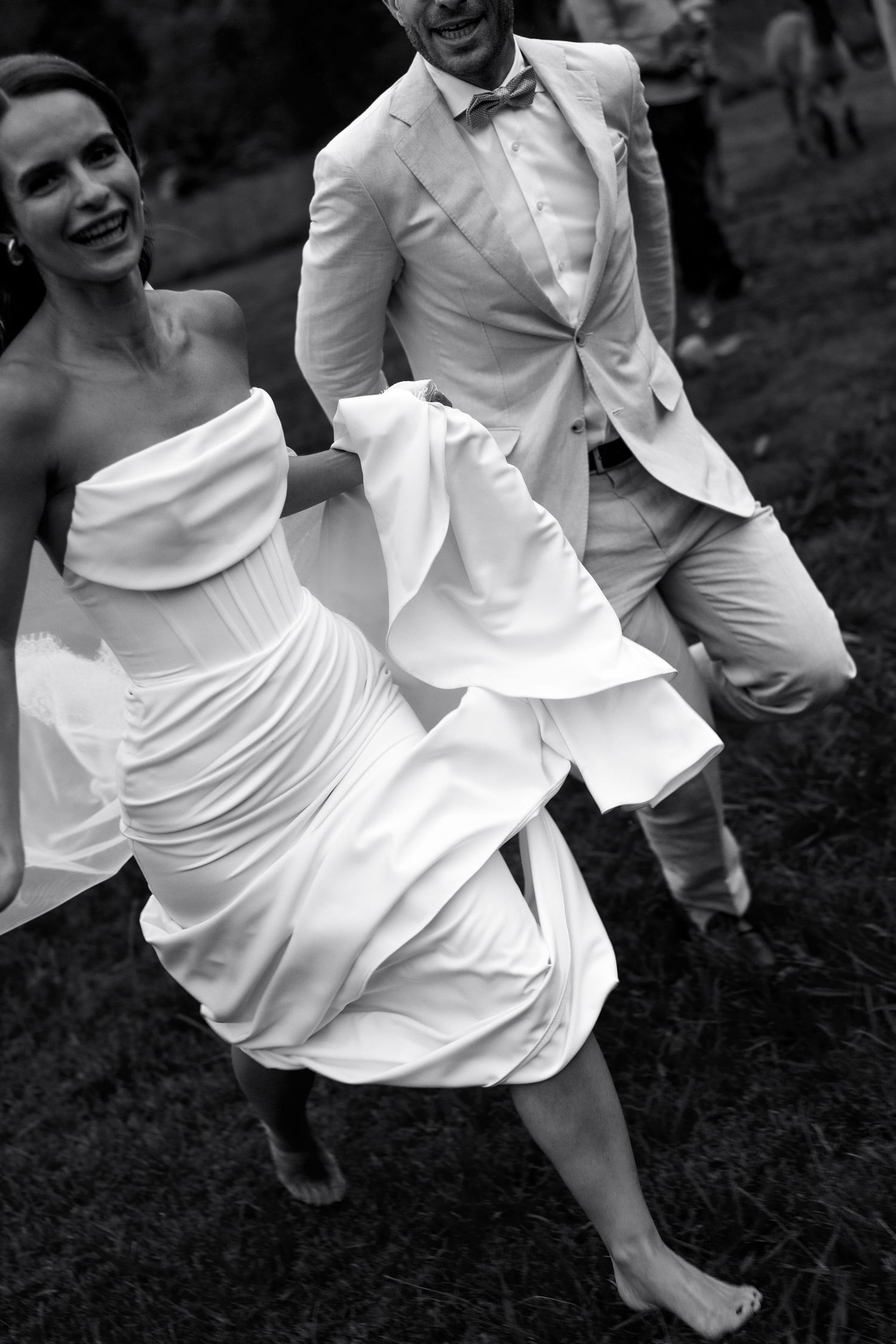 Black and white photo of a bride in a strapless dress twirling barefoot outdoors with a groom in white suit nearby.
