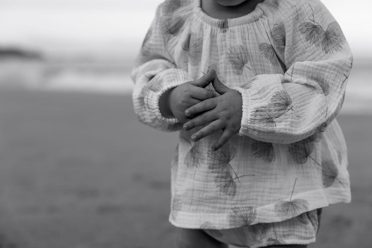 Black and white photo of a young child in a linen shirt clasping hands at the beach.