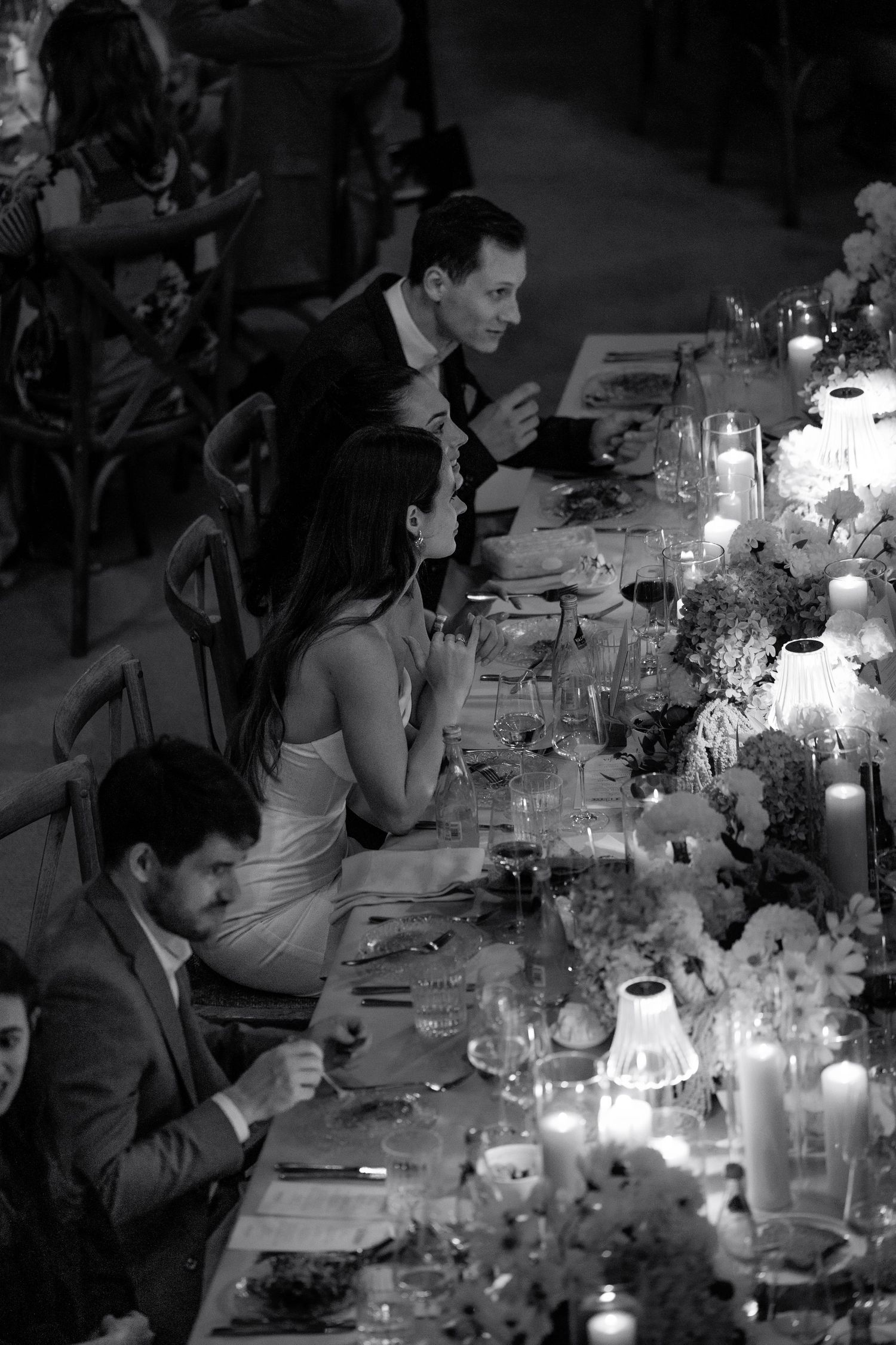 Black and white photo of elegantly dressed wedding guests dining at a candlelit table with floral arrangements.