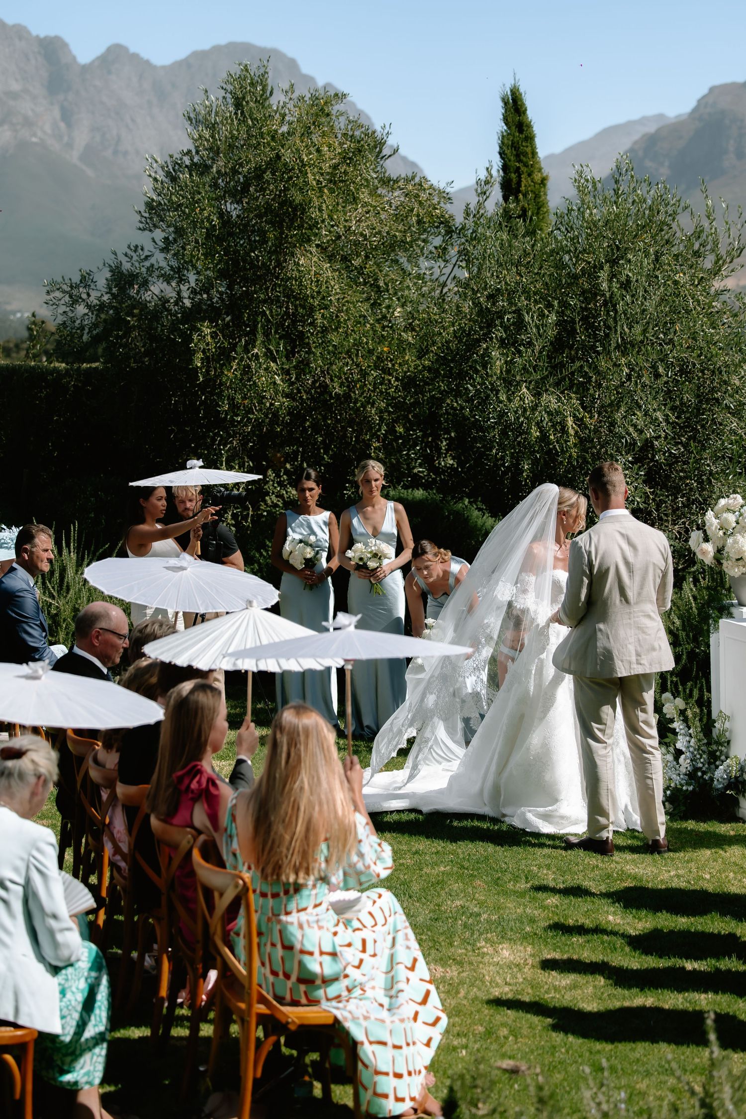 Outdoor garden wedding ceremony with white parasols and mountain backdrop.