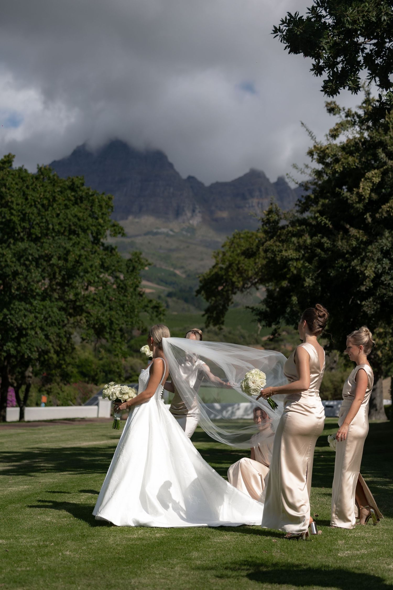 Bridesmaids help adjust a bride's flowing white dress train on a scenic mountain golf course with cloudy skies.