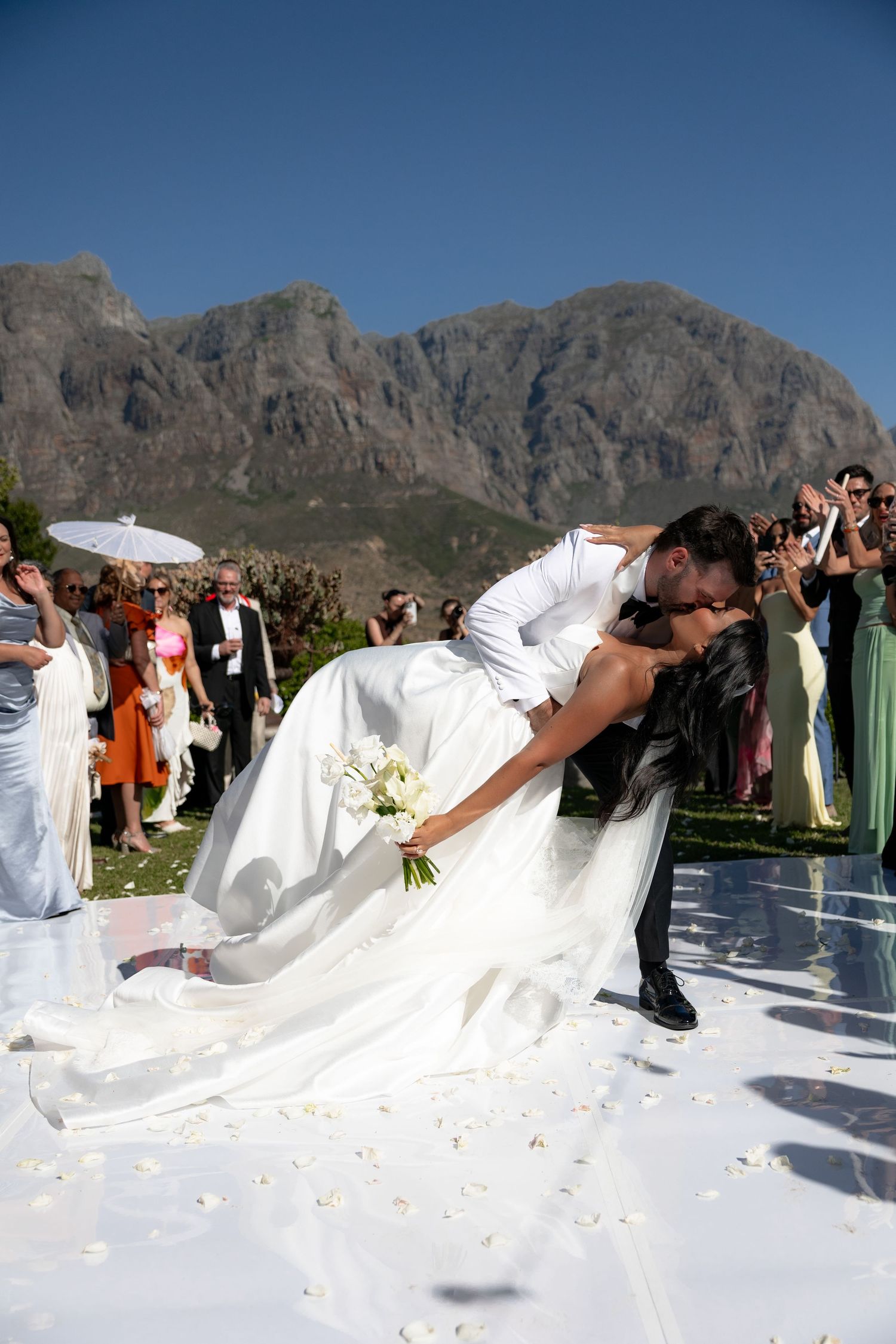 A wedding party poses with flowing white dresses against a dramatic mountain backdrop on a sunny day.