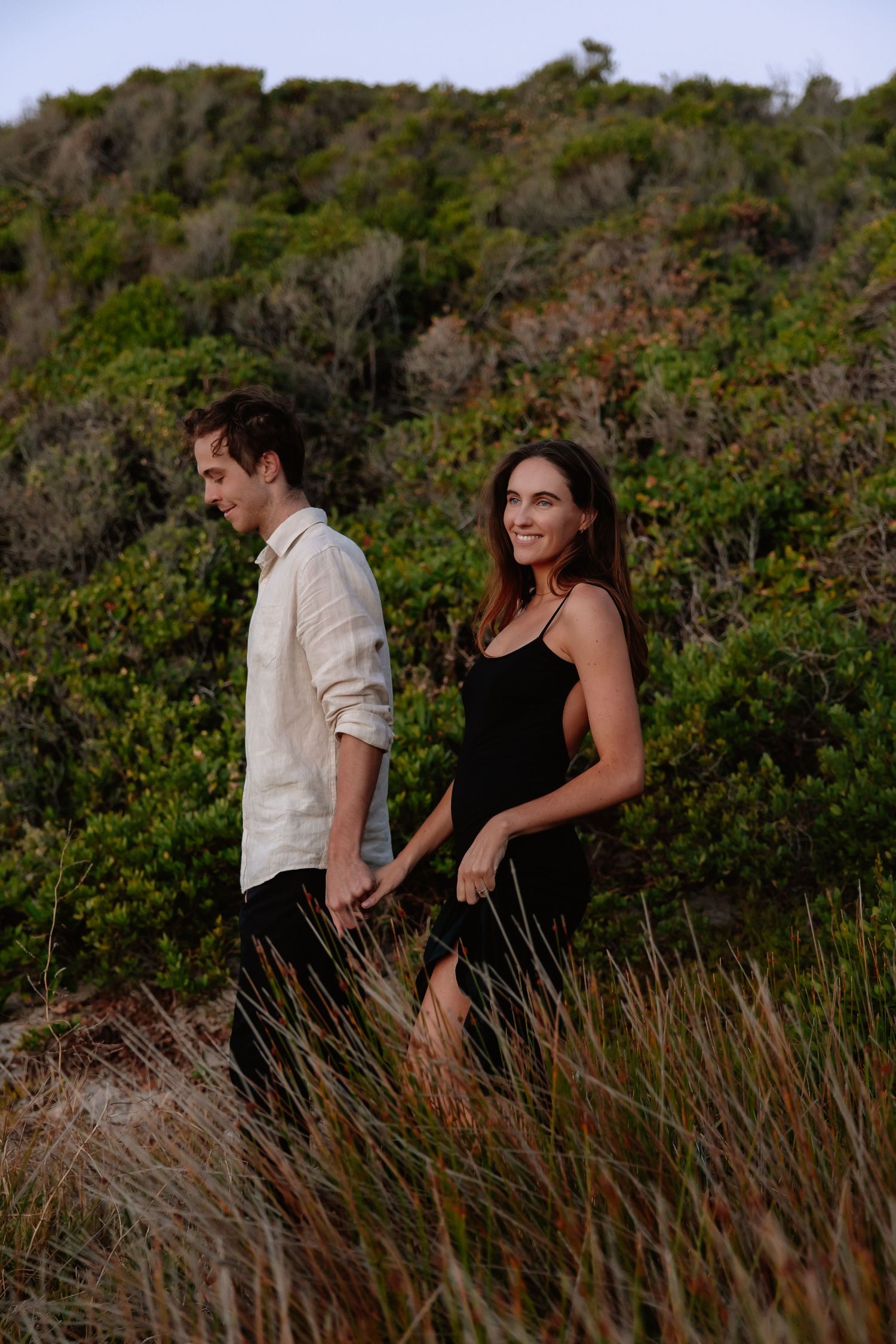 Couple holding hands walking through tall grass with lush green bushes, woman in black dress, man in white linen shirt.