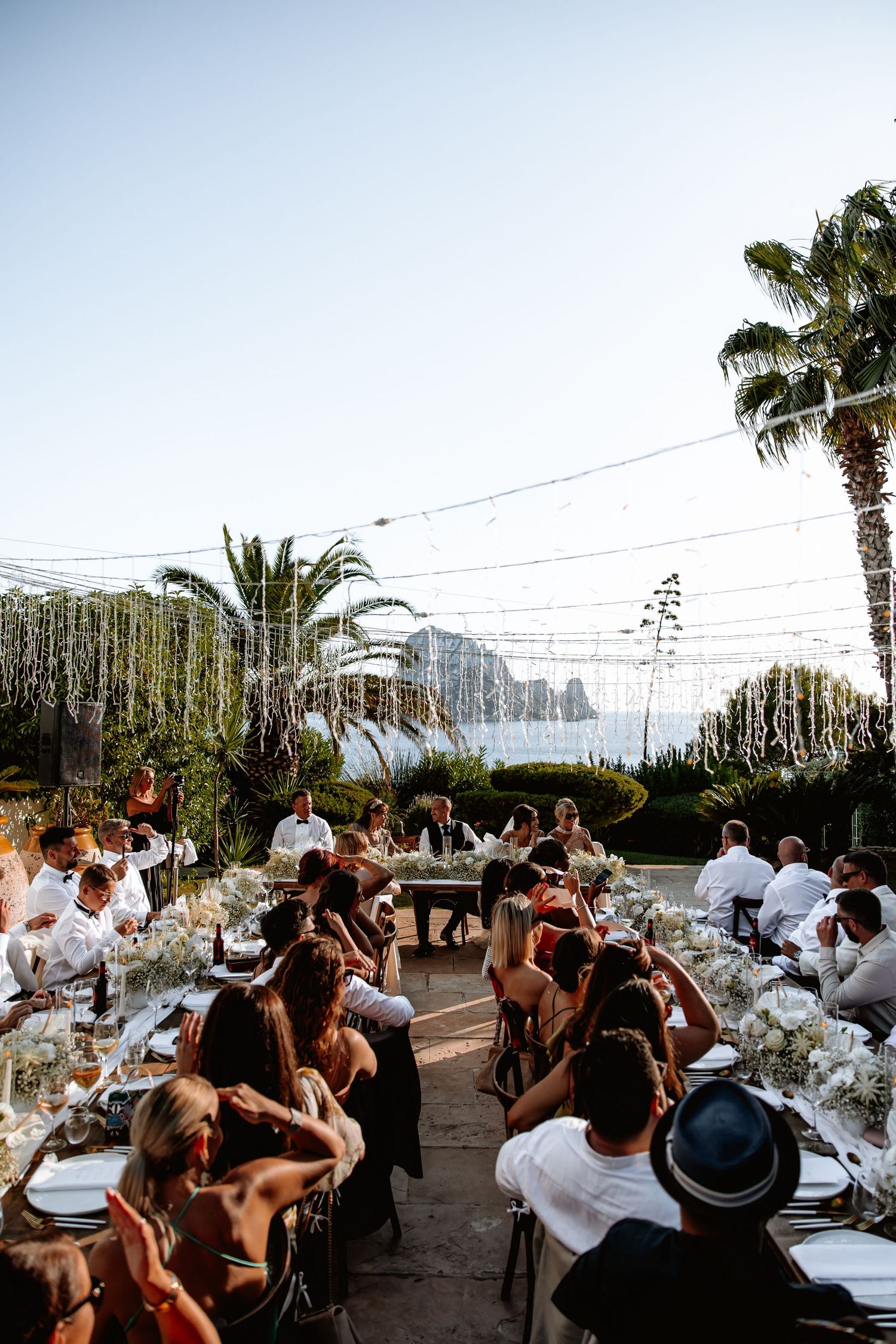 Wedding guests enjoy an outdoor reception dinner under palm trees and string lights at sunset.