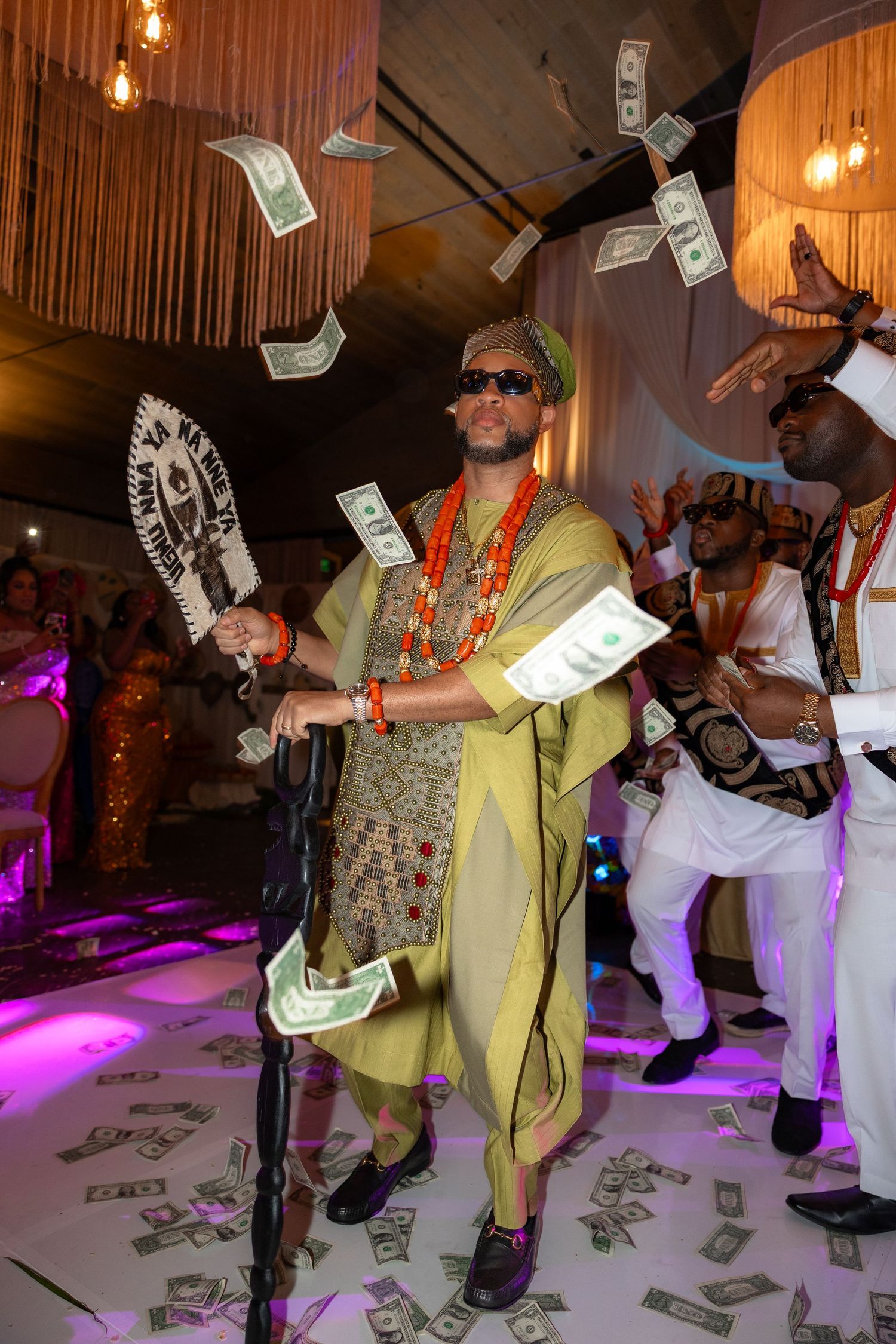 Money rains down on a dance floor as people celebrate under warm lighting and purple accents.