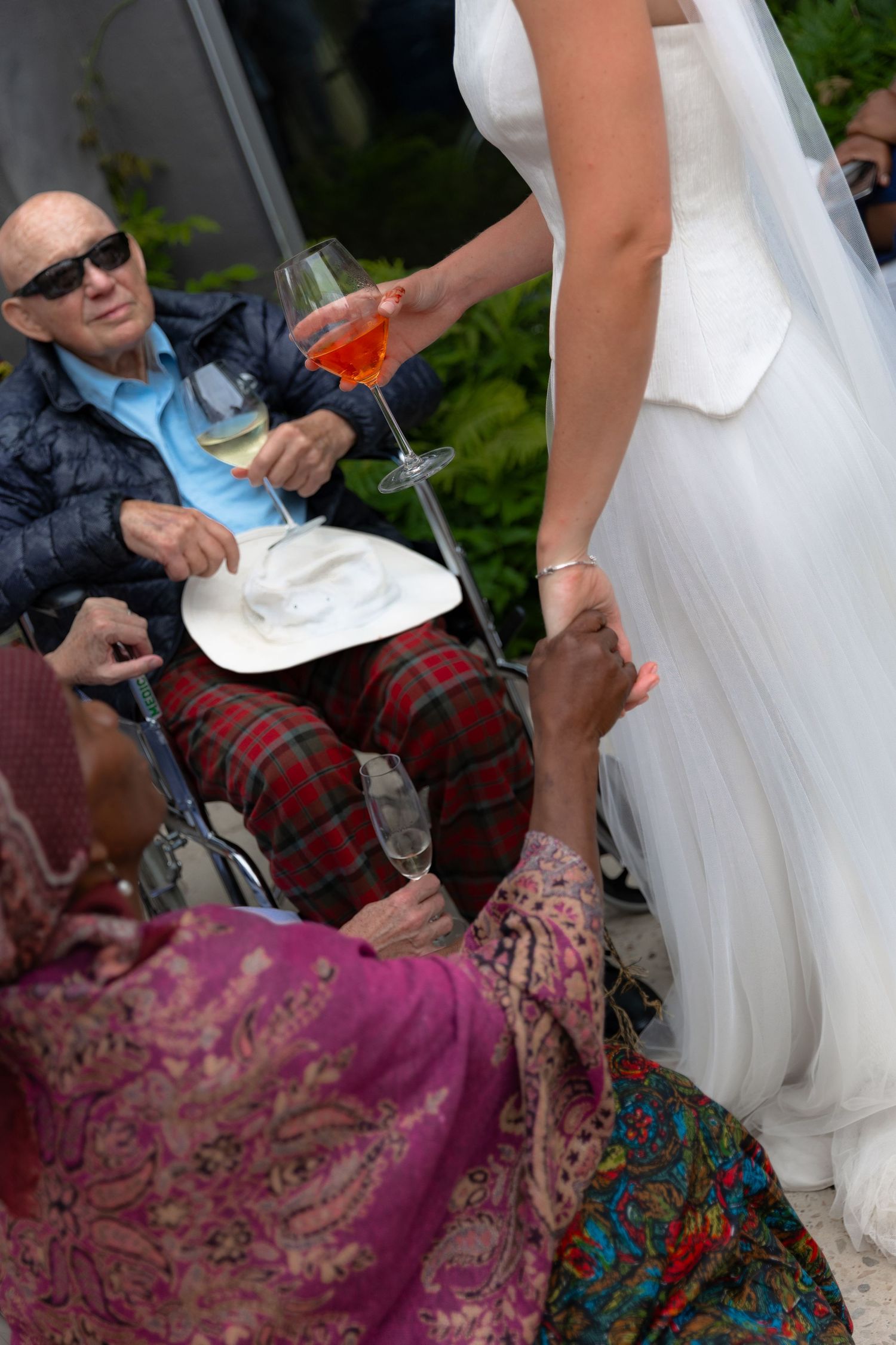 Bride in white gown holds hands with elderly man in wheelchair at outdoor wedding reception with drinks.