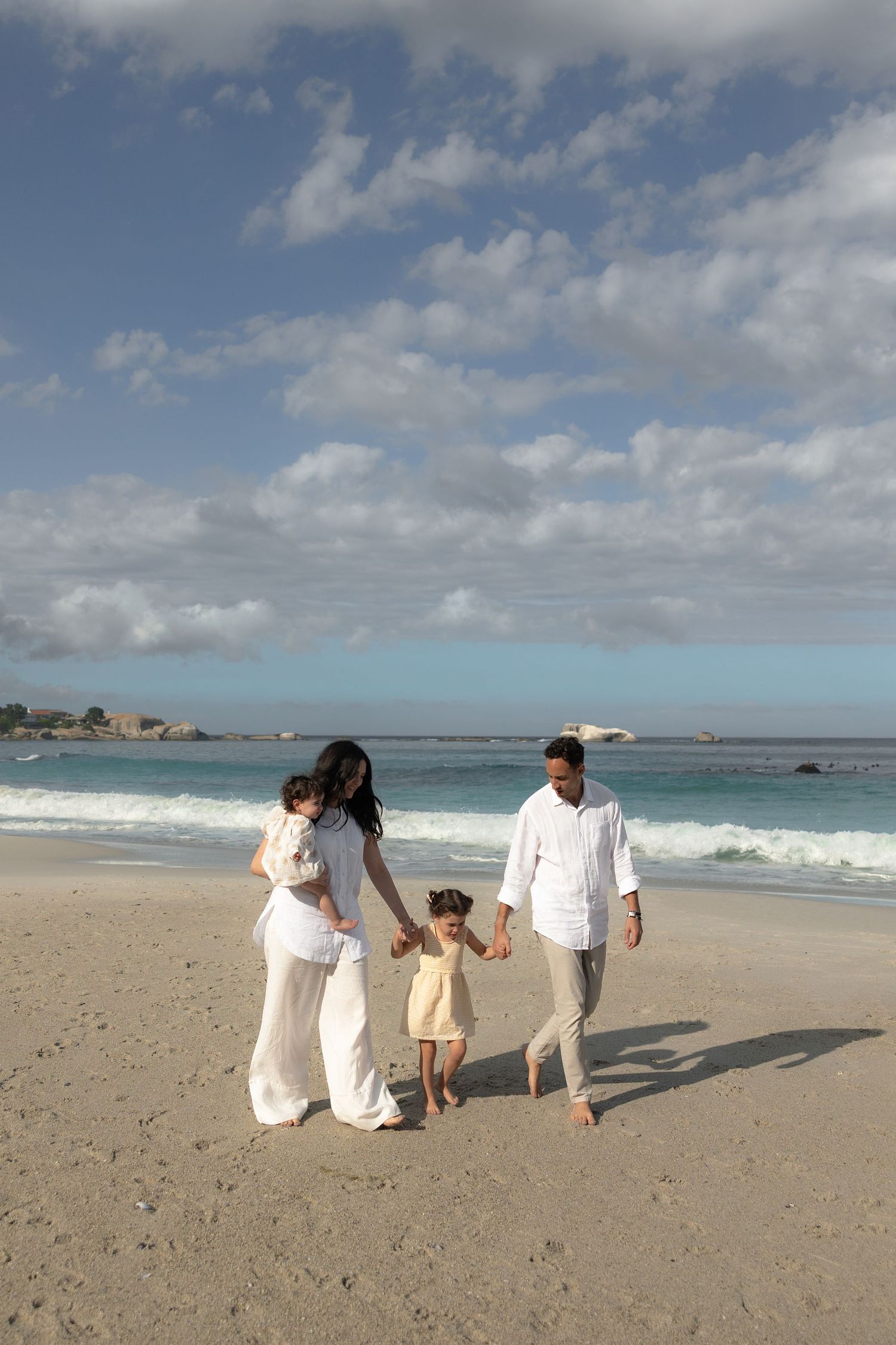 Family in white outfits walking together on a sunny beach with ocean waves and blue sky in the background.