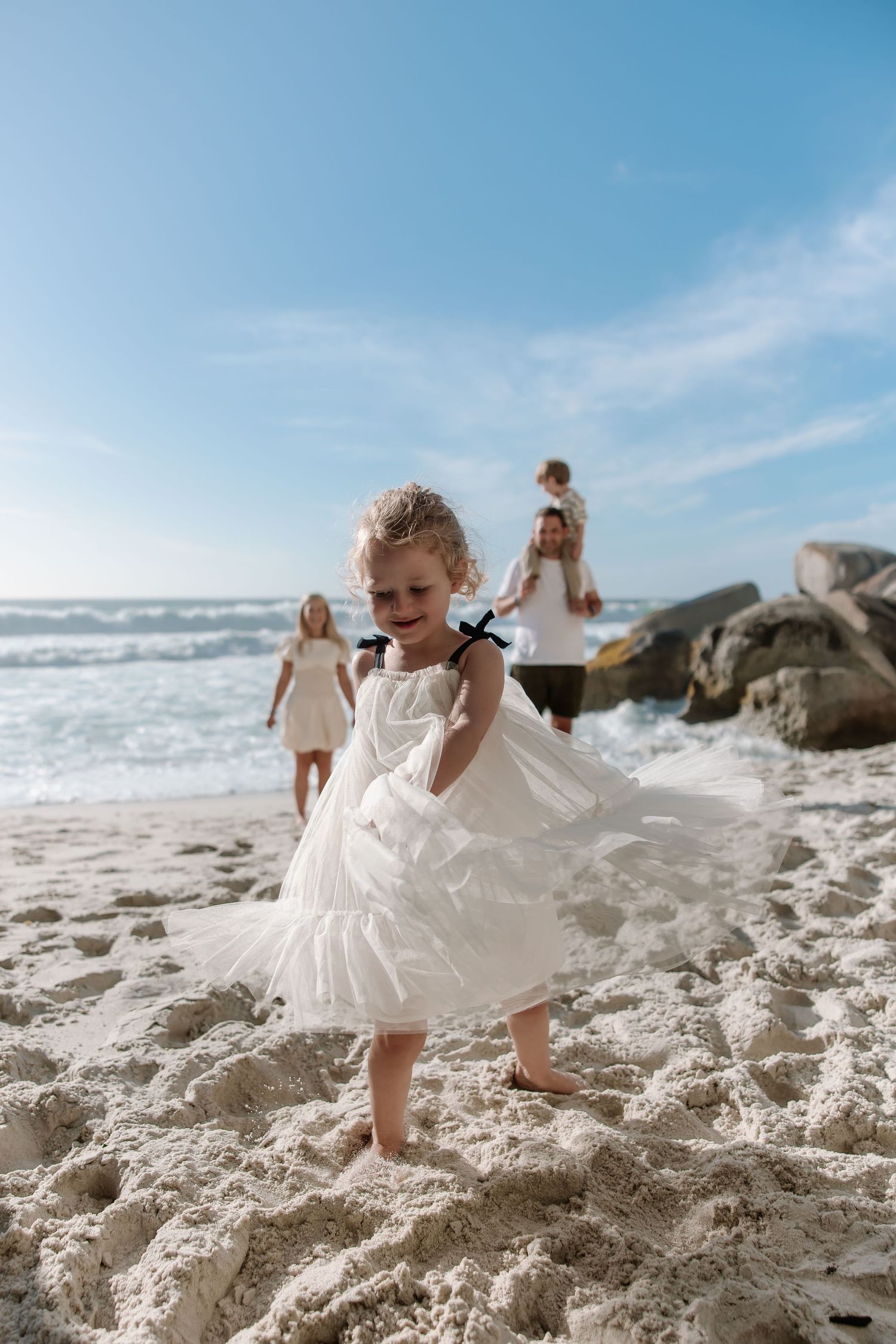 A child in a white dress twirls on a sunny beach with waves and rocks in the background.