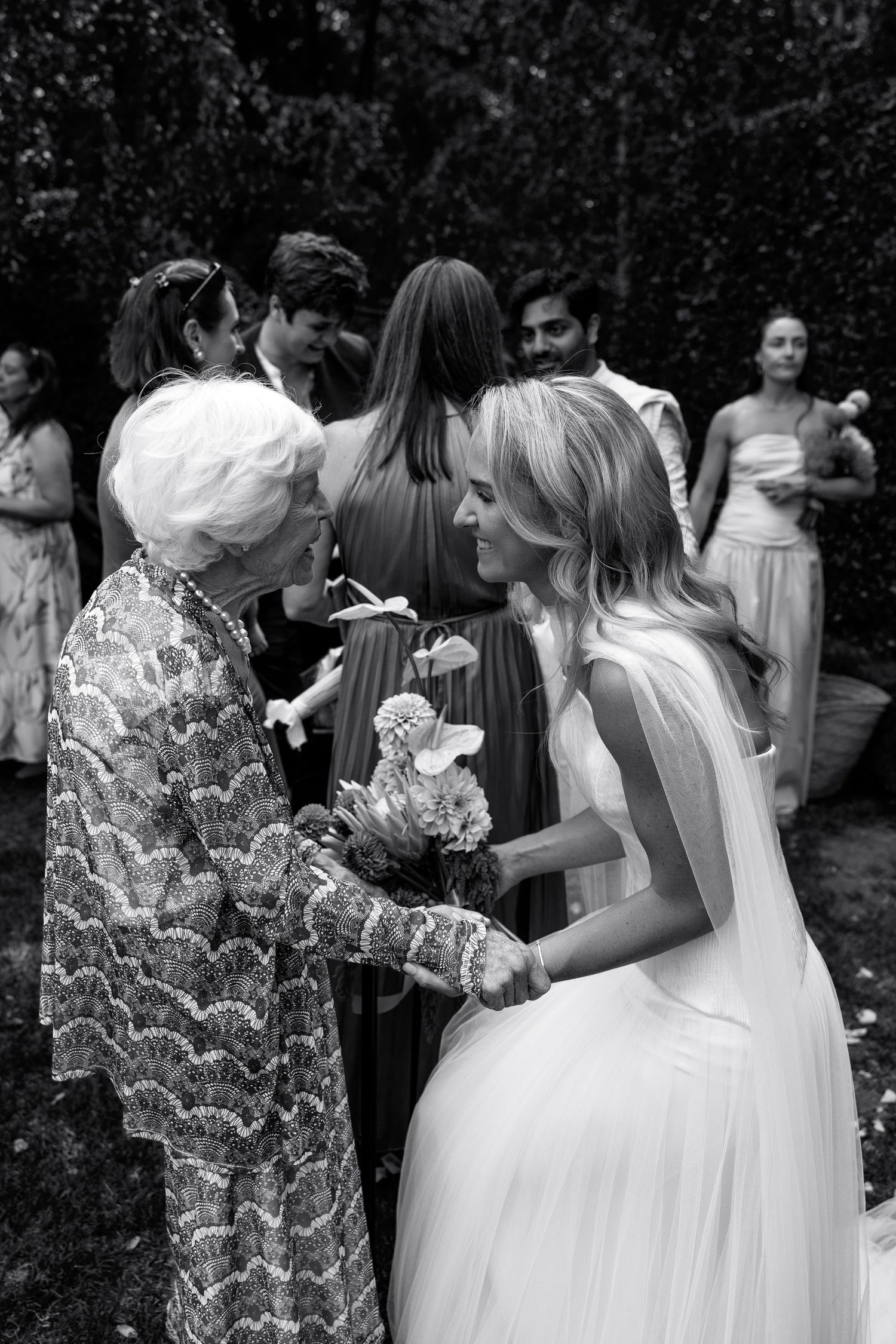 Black and white photo of a bride in white gown receiving congratulations from an elderly woman at an outdoor wedding.