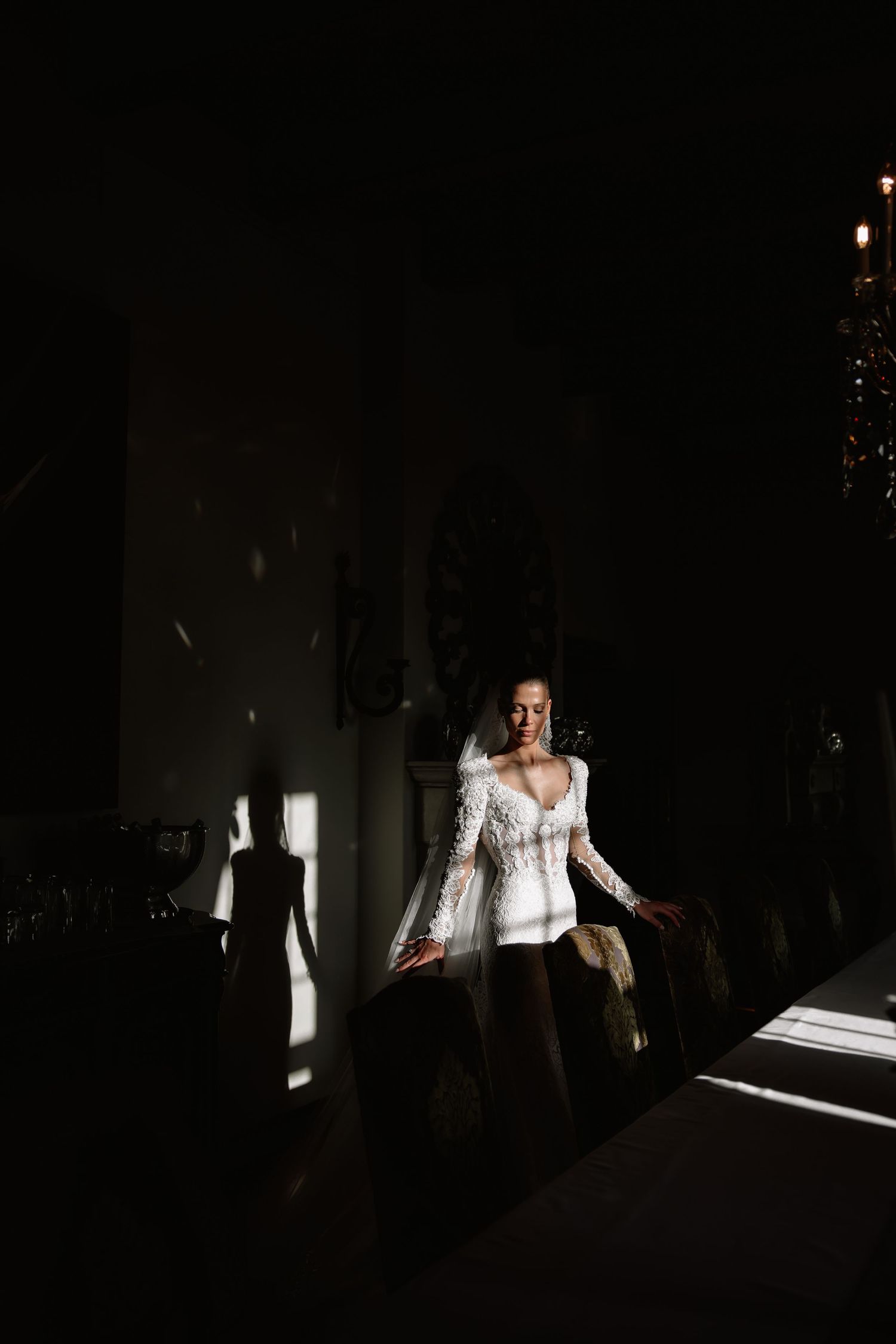 Dramatic bridal portrait in a dark room with light streaming through windows highlighting a white lace gown.
