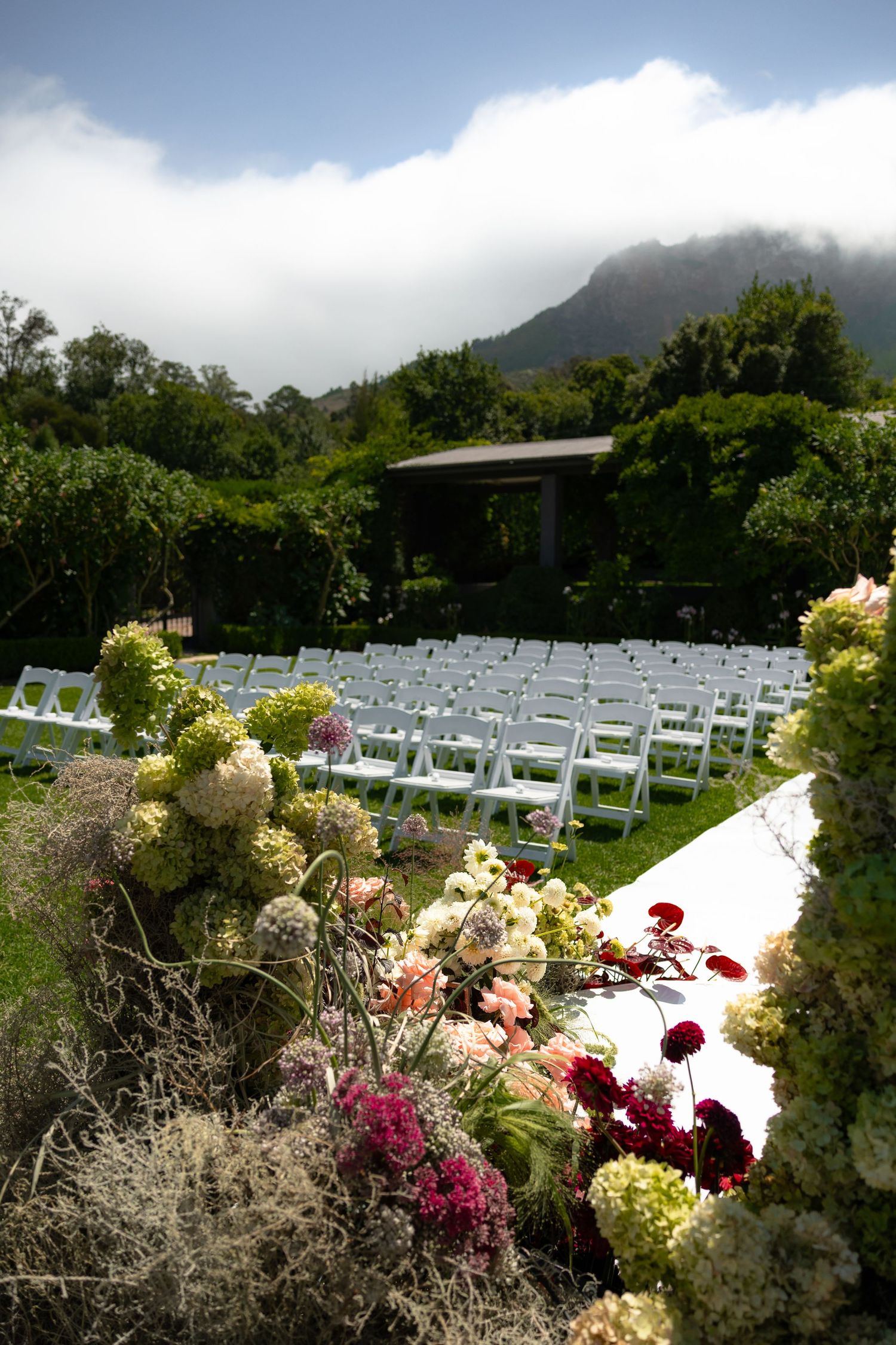White chairs arranged for outdoor wedding ceremony with mountain backdrop and colorful floral garden border.