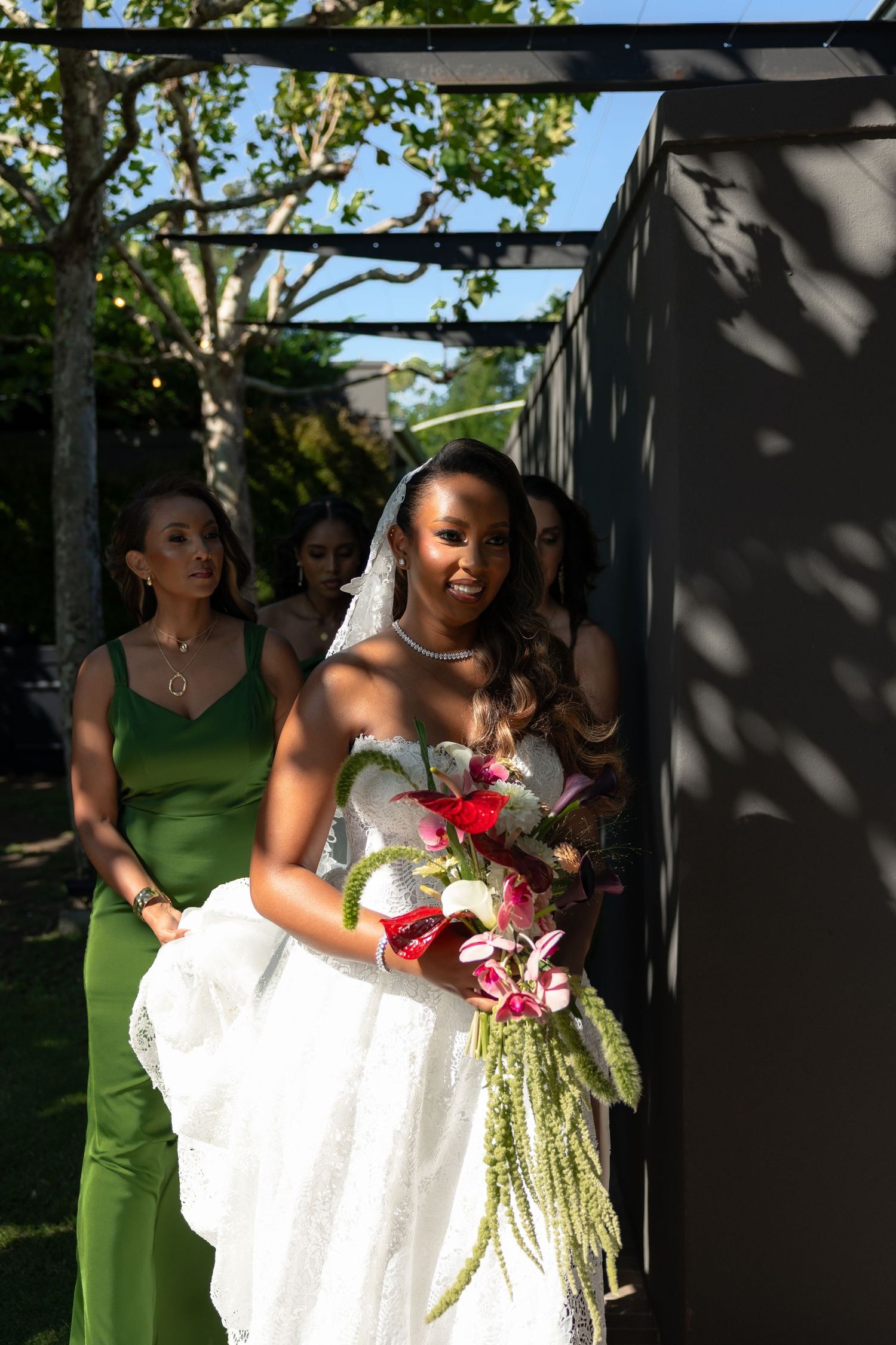 Wedding party in green dresses and white gown pose together outdoors under tree canopy with floral bouquets.