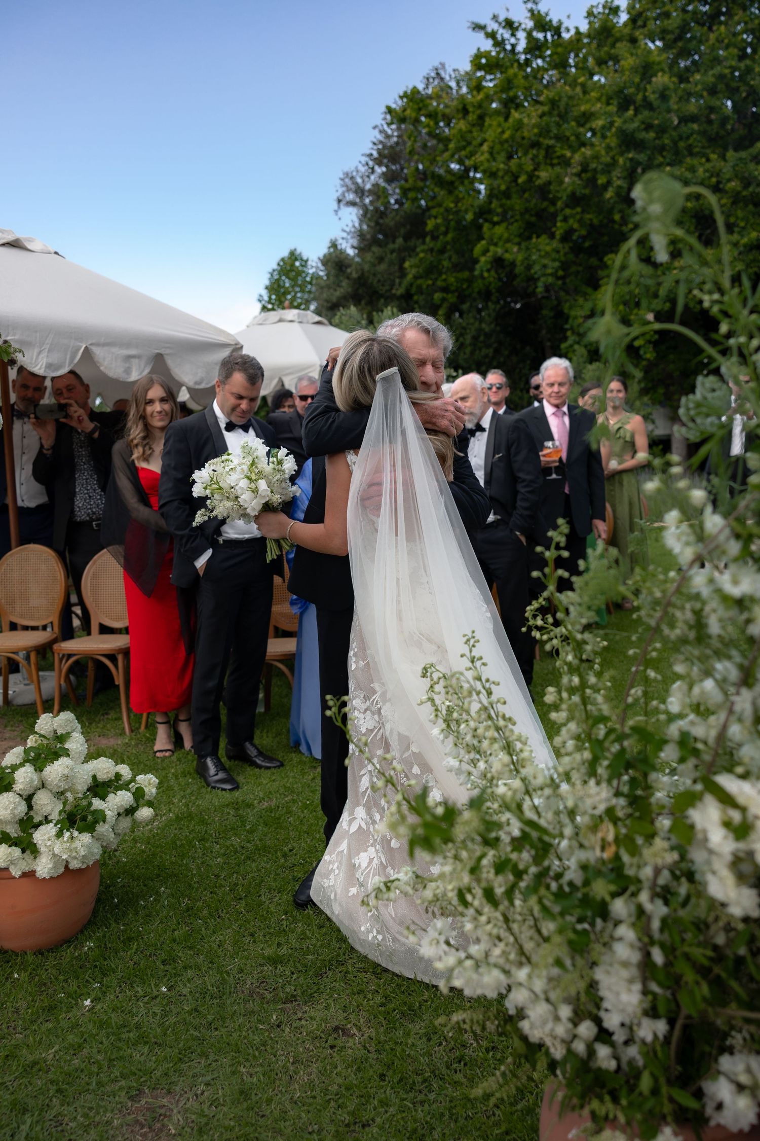 Wedding guests gather outdoors on a lawn while a long white veil flows in the breeze among blooming white flowers.