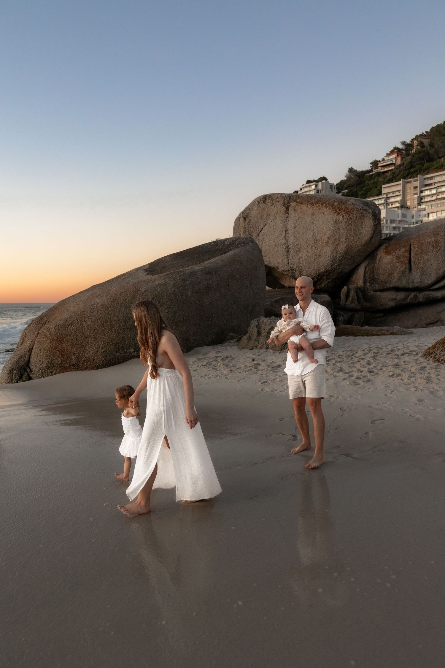 Family in white outfits enjoying a sunset beach walk, with large rocks and ocean waves in the background.