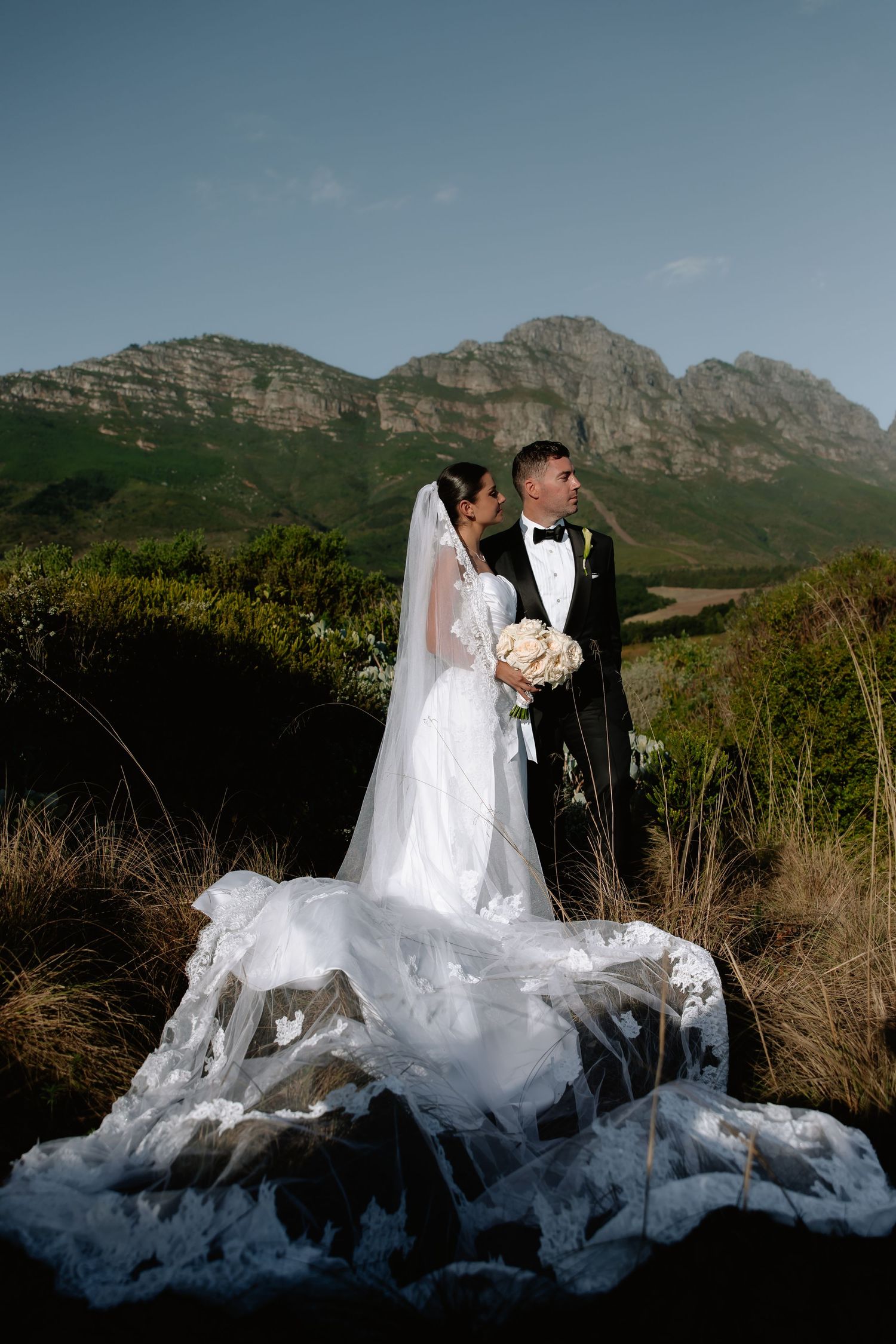 Wedding couple poses against dramatic mountain backdrop at sunset with flowing white dress creating ethereal silhouette.