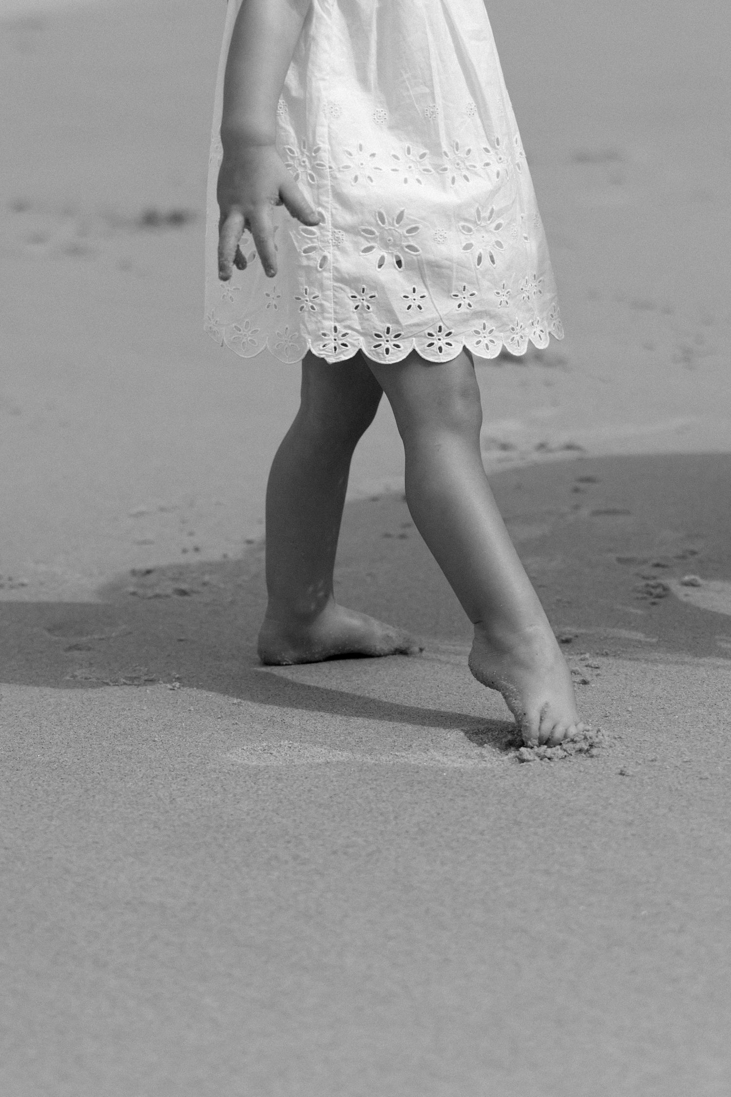 Black and white close-up of small feet in sand wearing white eyelet dress.