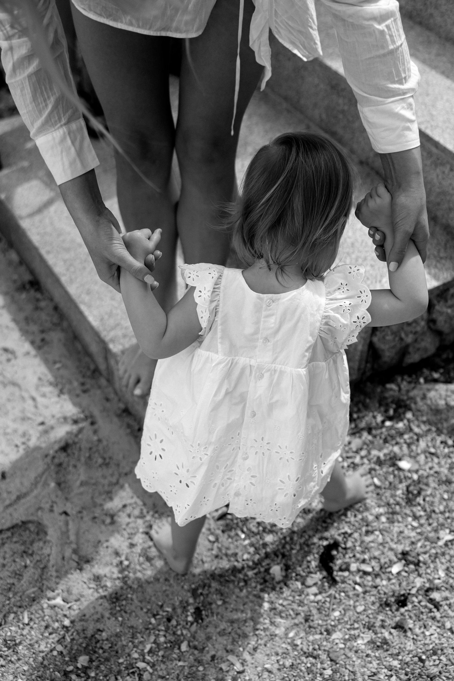 Black and white image of small child in white eyelet dress holding hands with adults.