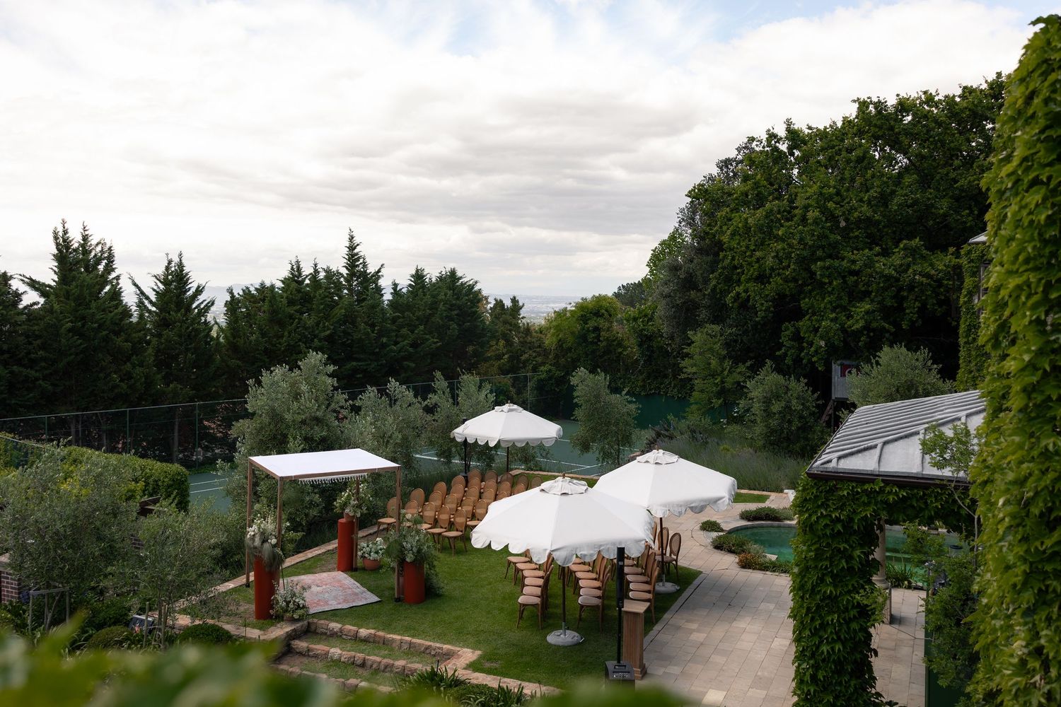 Outdoor garden venue with white umbrellas and seating areas surrounded by lush greenery and trees.