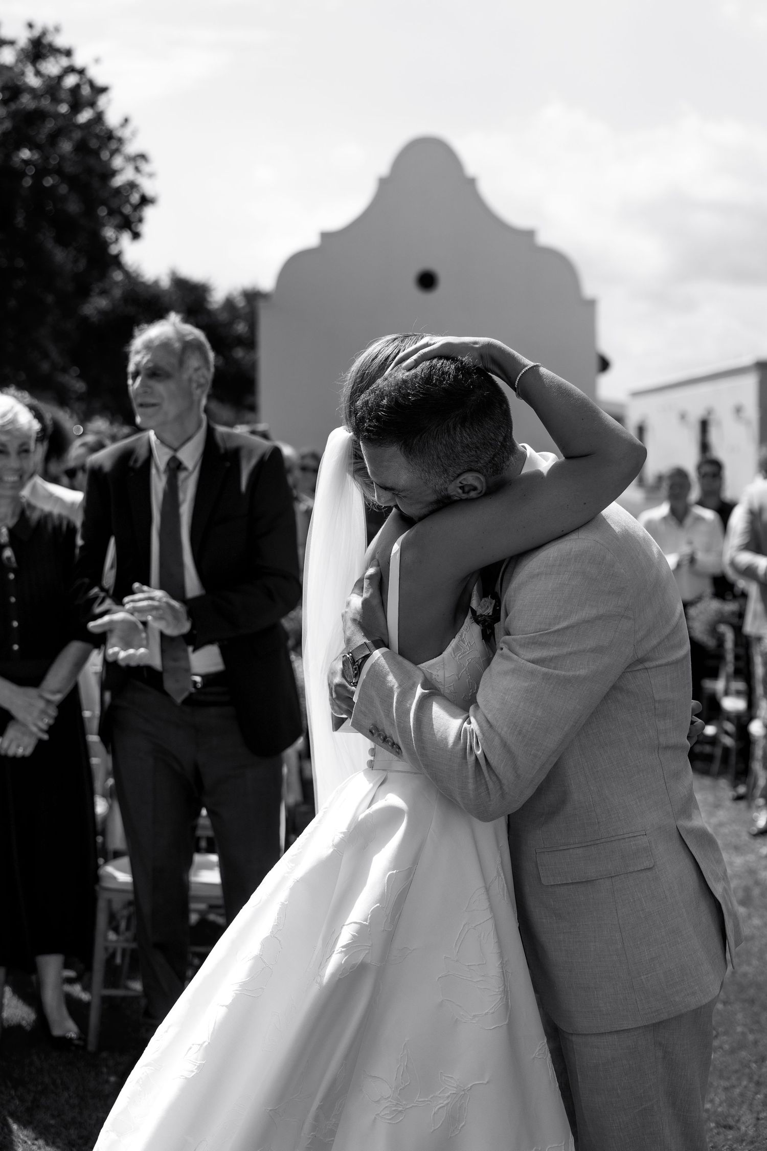 Black and white wedding photo showing emotional embraces between wedding party members outdoors.