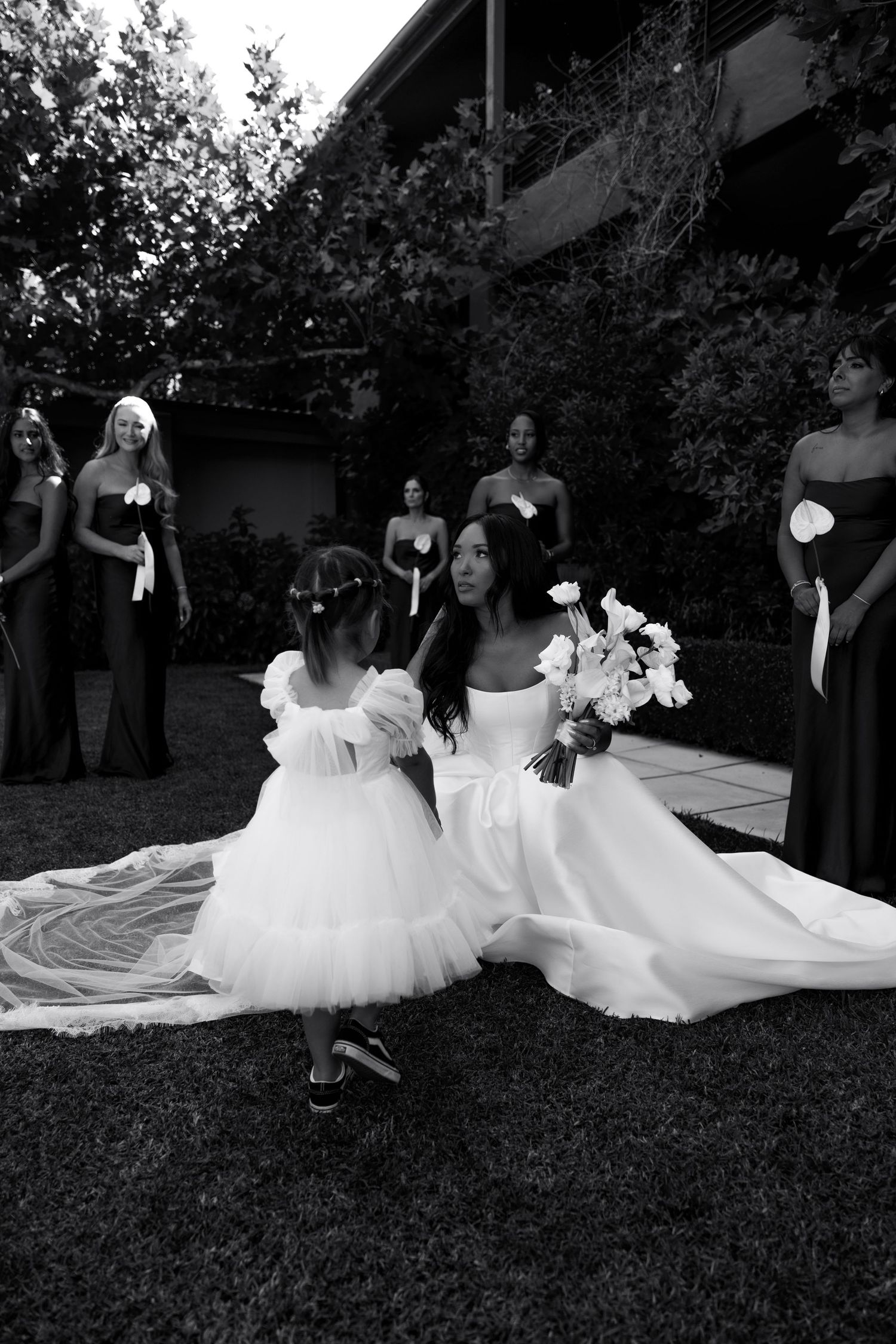 A black and white wedding photo showing a bride in a flowing gown with attendants in dark dresses on a lawn.