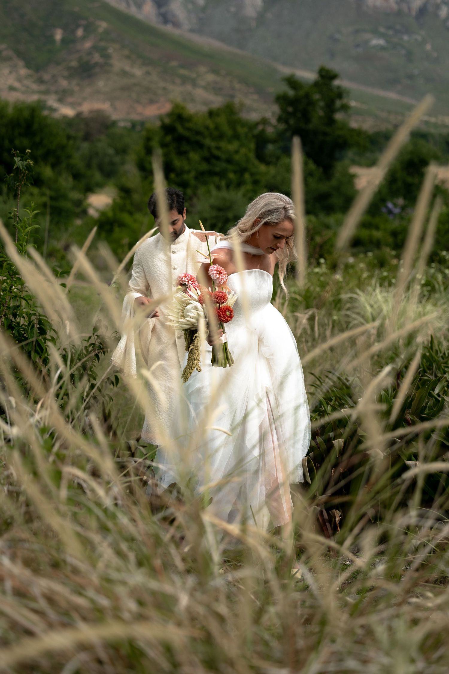 Couple in white wedding attire share romantic moments in a rustic field with tall grass and mountains in background.