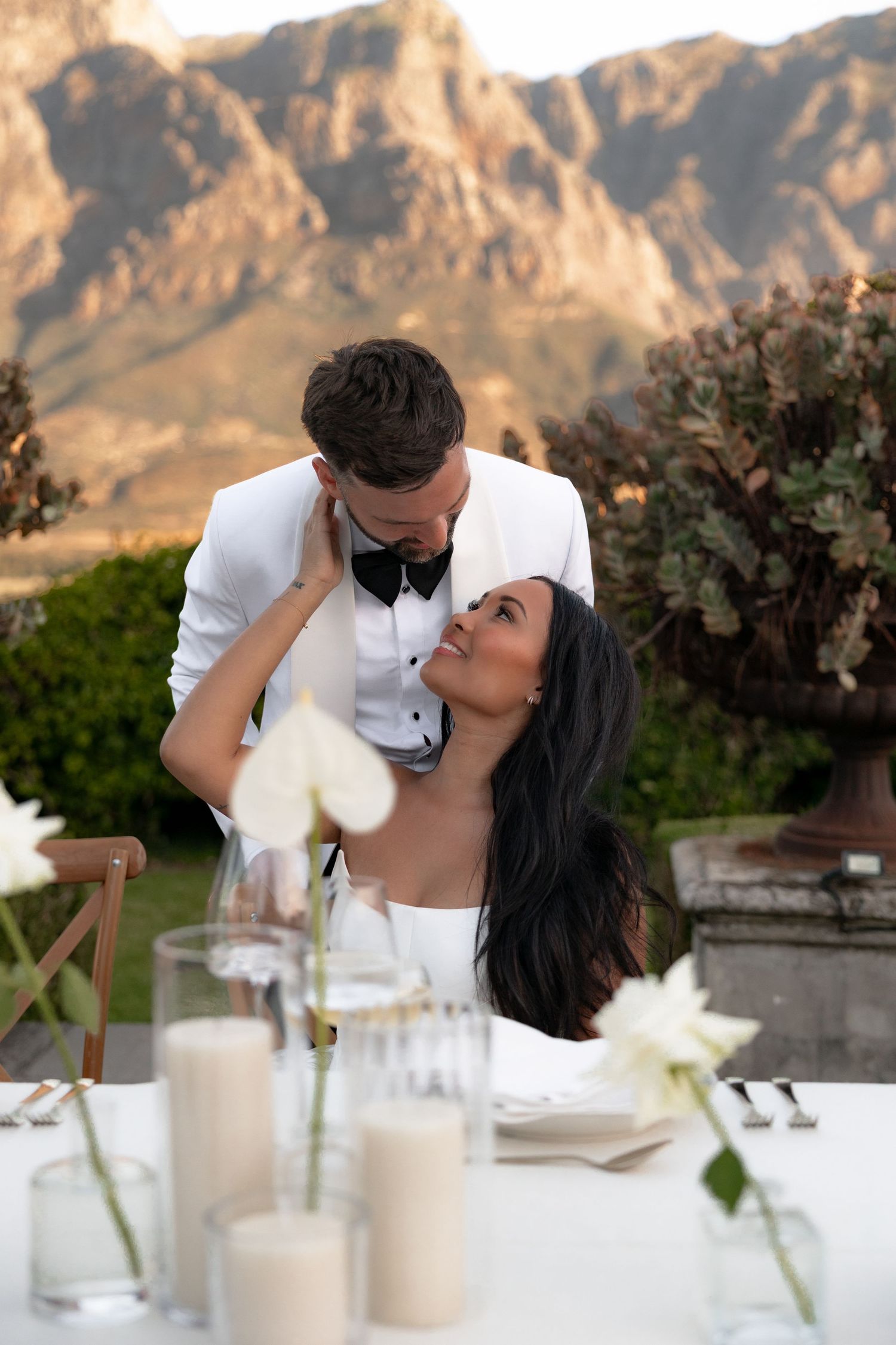 Romantic wedding couple sharing an intimate moment at an outdoor reception with mountain backdrop and candlelit table.