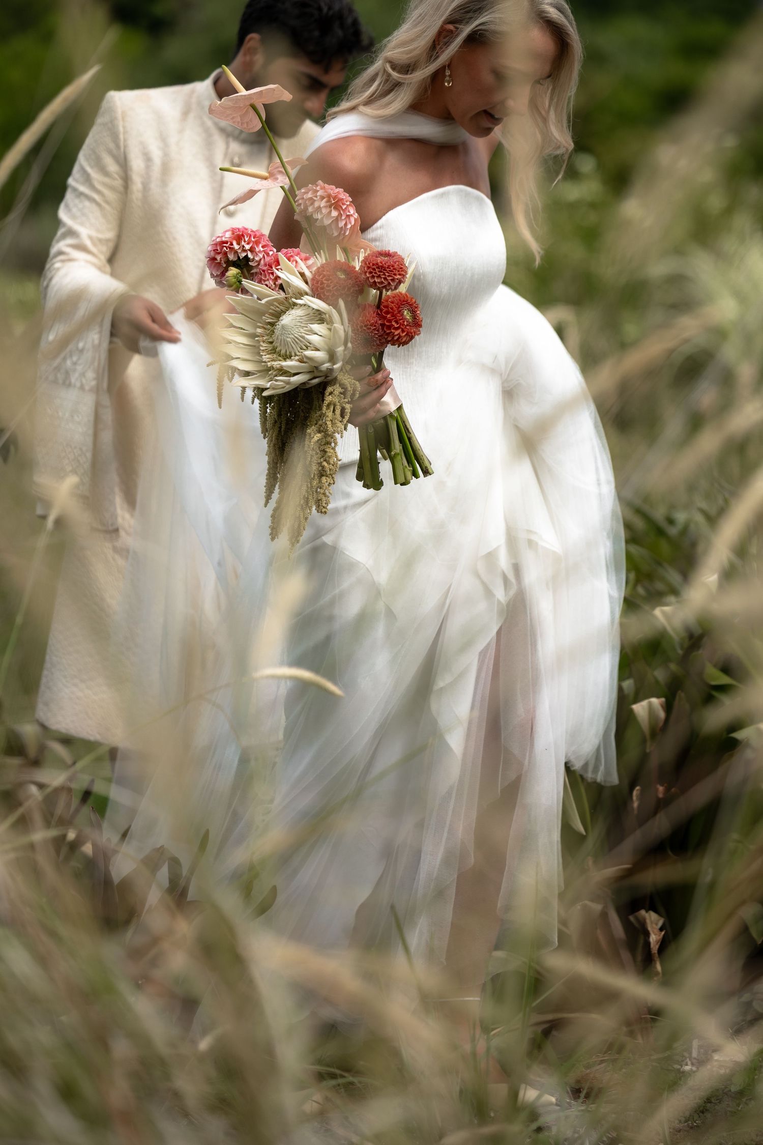 Artistic wedding photo through foliage showing white dress details and red bouquet.