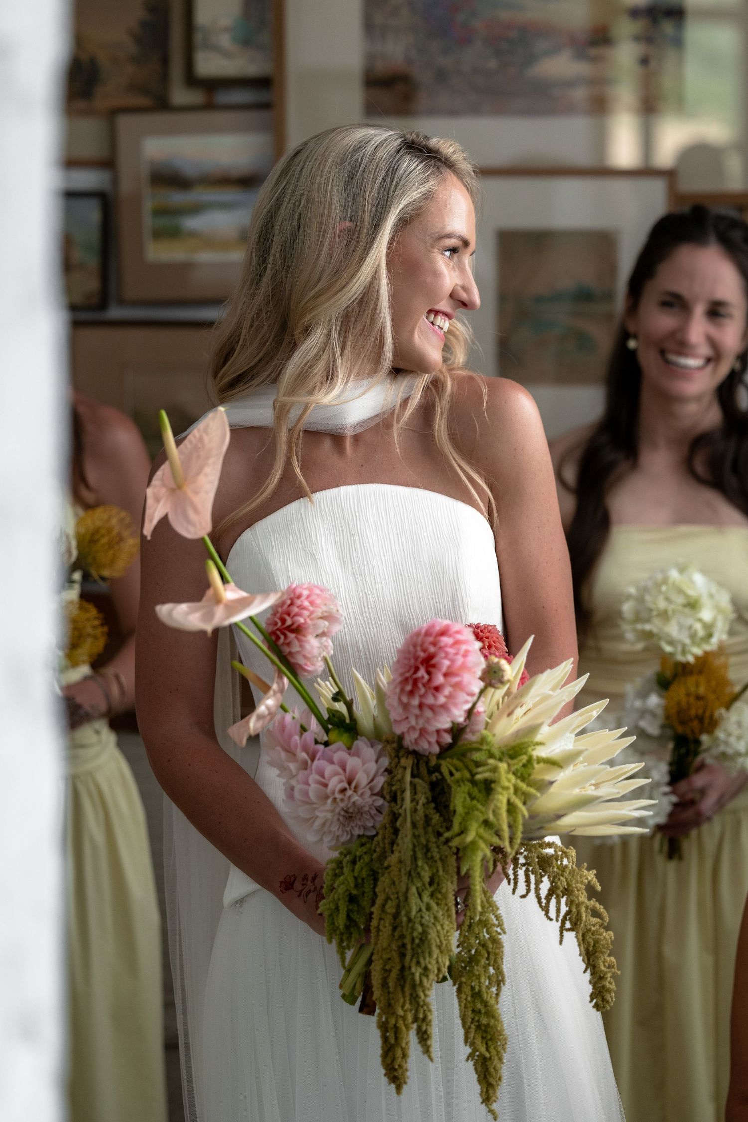 A bride in a white strapless gown holds a beautiful bouquet of pink dahlias and greenery while smiling during a wedding.