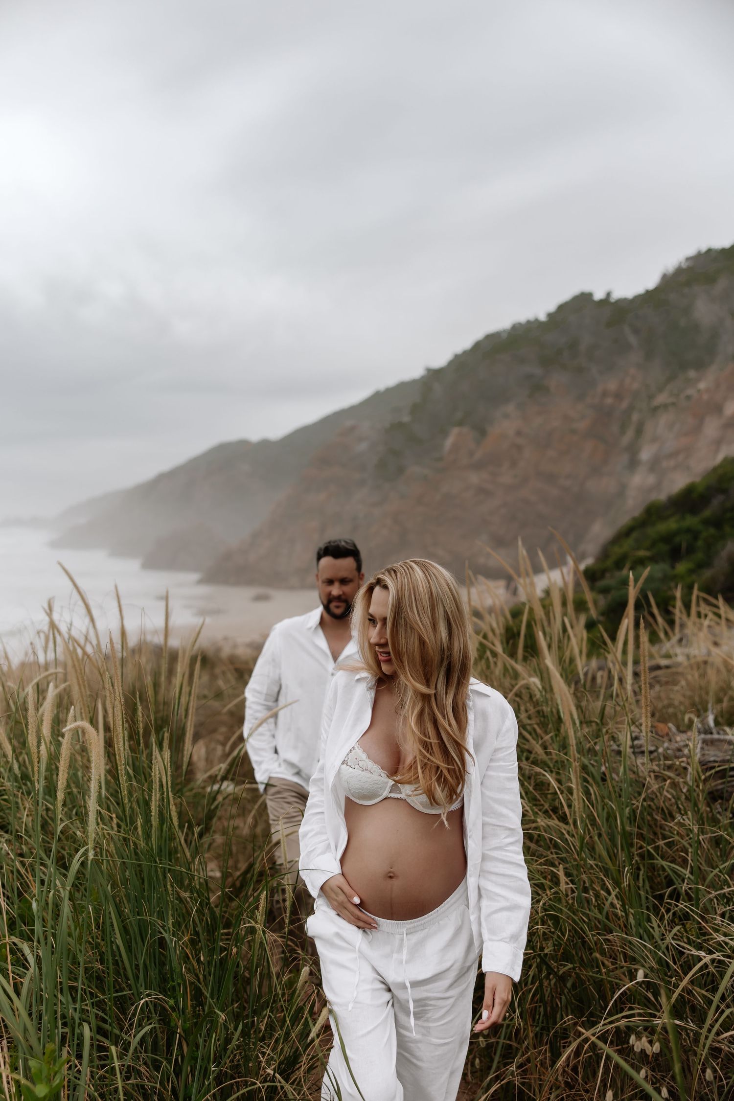 Romantic coastal photoshoot with couple in white outfits standing among tall grass with foggy cliffs in background.