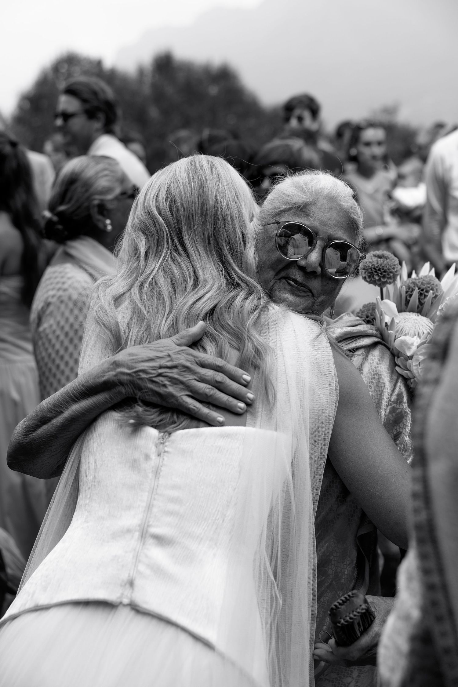 Black and white photo of a bride hugging an elderly woman wearing large glasses at an outdoor wedding celebration.