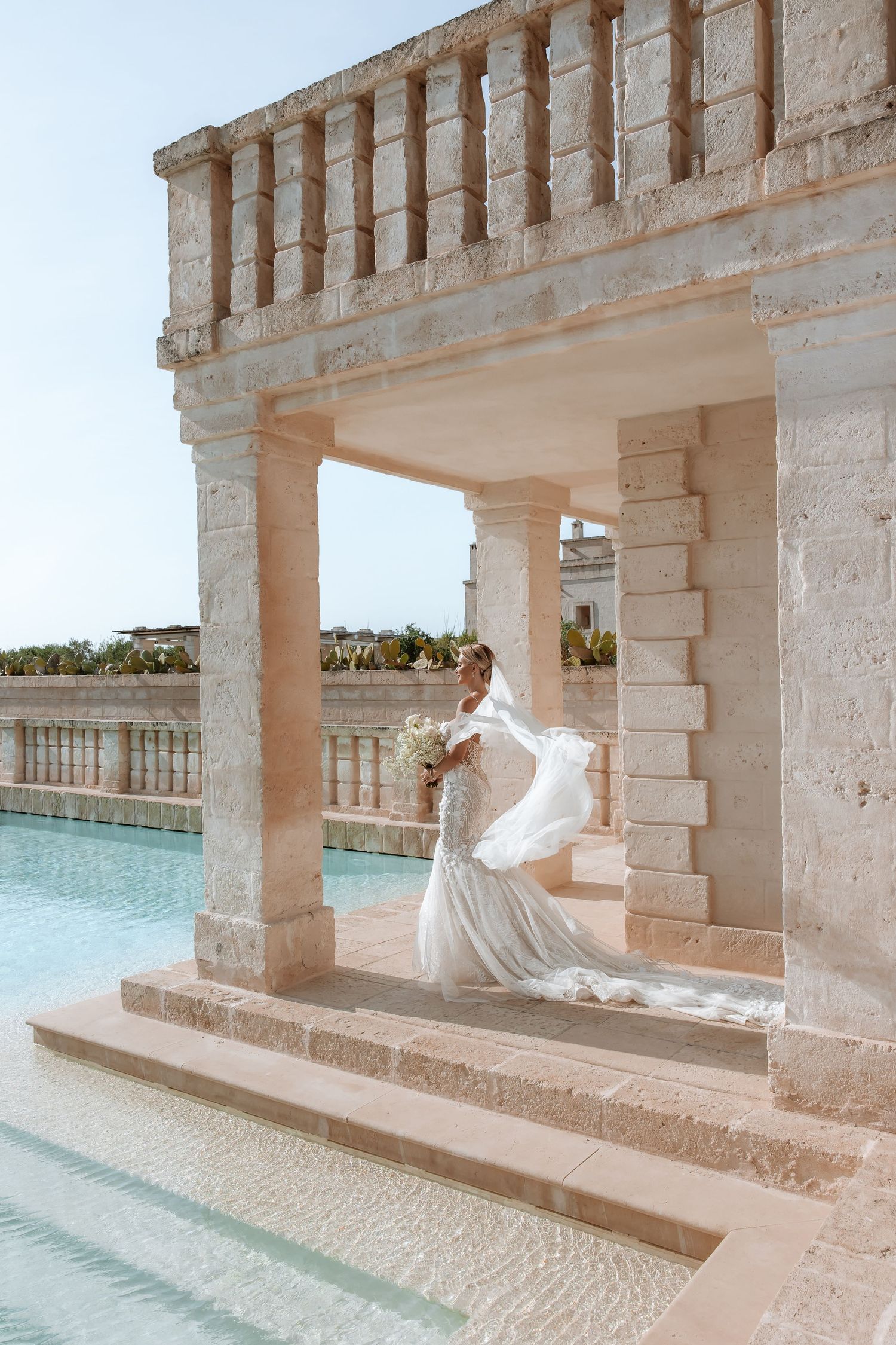 Wedding dress with long train photographed under stone colonnade beside turquoise pool.