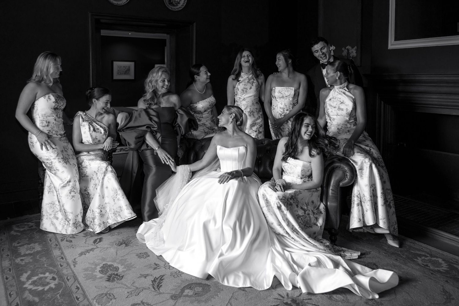 Black and white photo of a bride in a white gown laughing joyfully with her bridesmaids in elegant dresses.