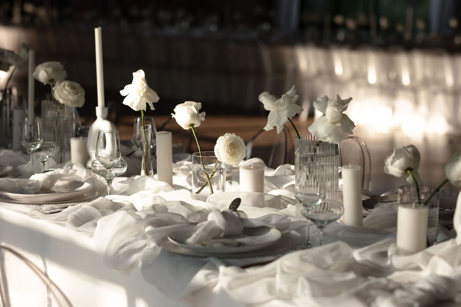 Elegant white wedding reception table with candles and floral centerpieces in sunlight.