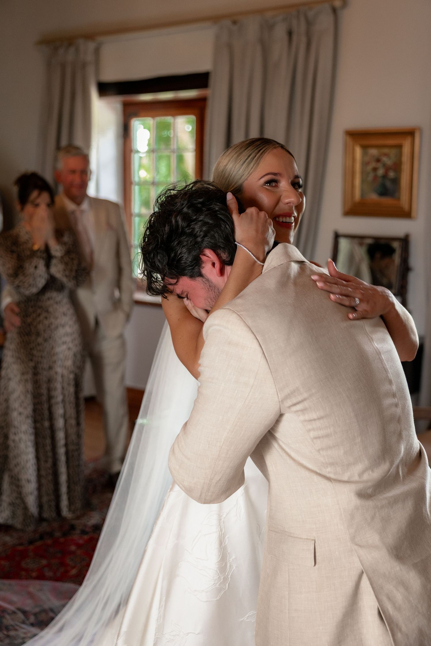A tender embrace between a couple at a wedding celebration, with guests visible in the background.