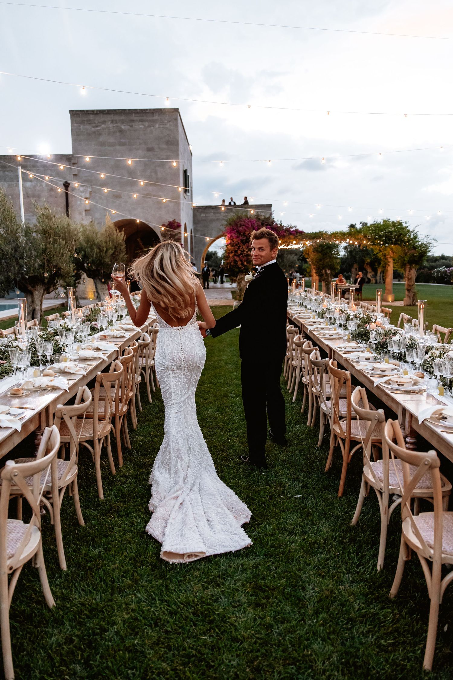 Elegant outdoor wedding reception setup with long tables, string lights, and white chairs on a grassy lawn.