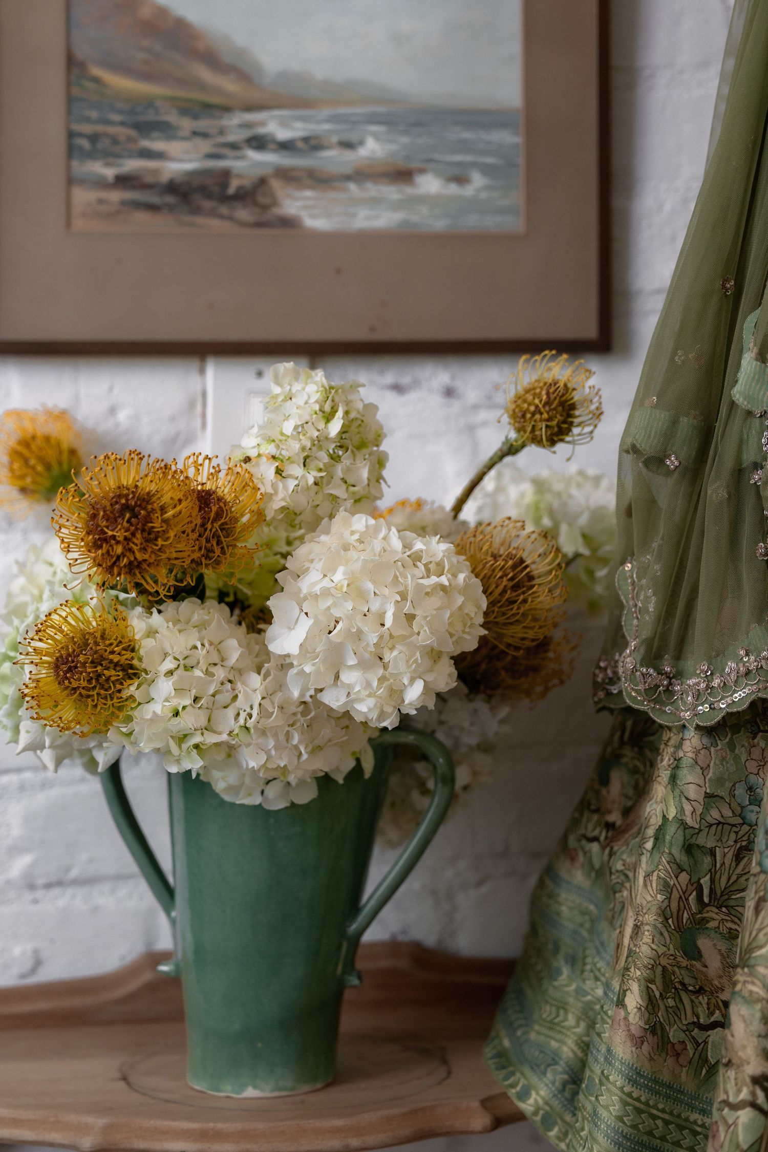 White hydrangeas and yellow flowers arranged in turquoise vases against a coastal artwork backdrop.